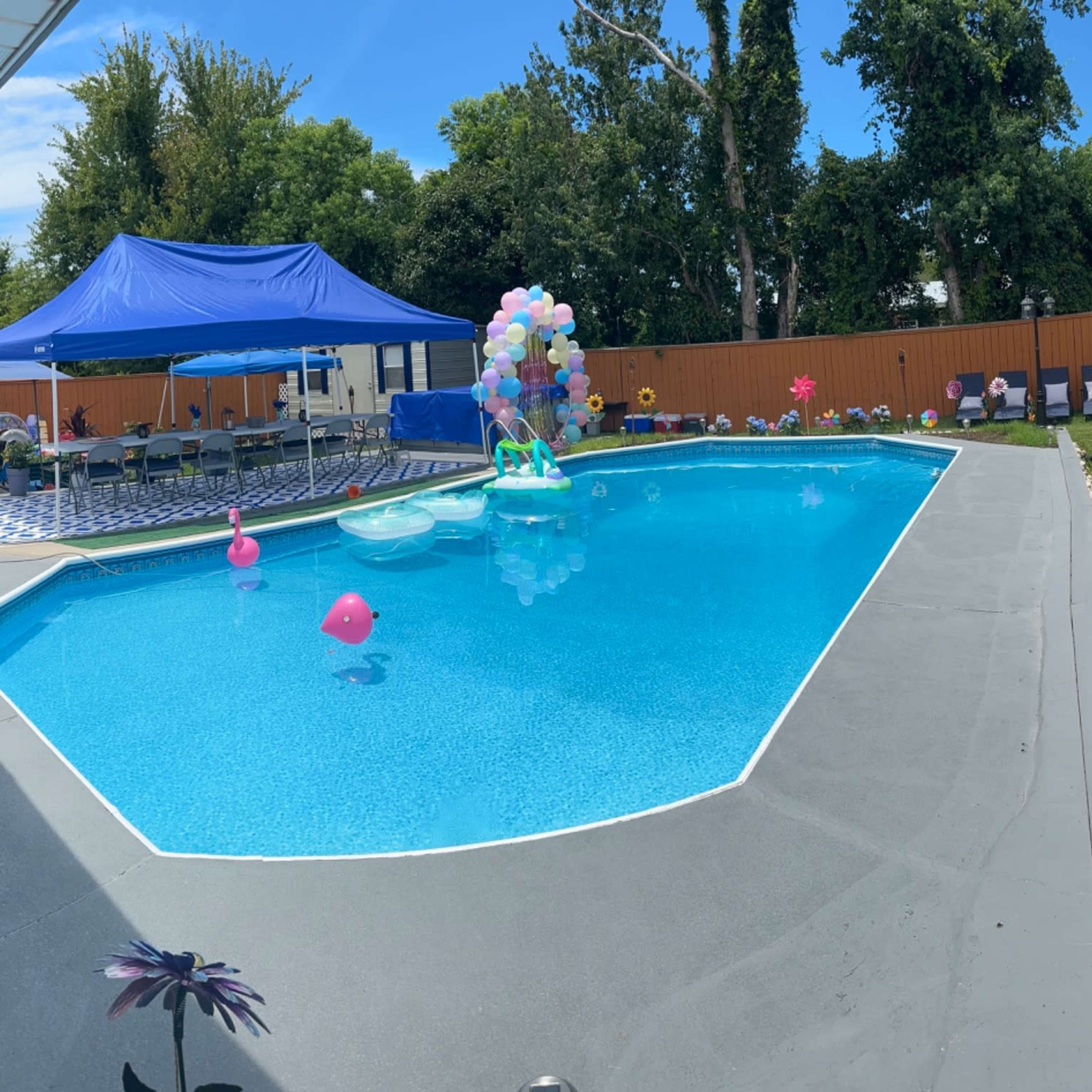 A panoramic view of a backyard pool area with a blue pool, lounge chairs, a tent, and colorful decorations around the poolside.