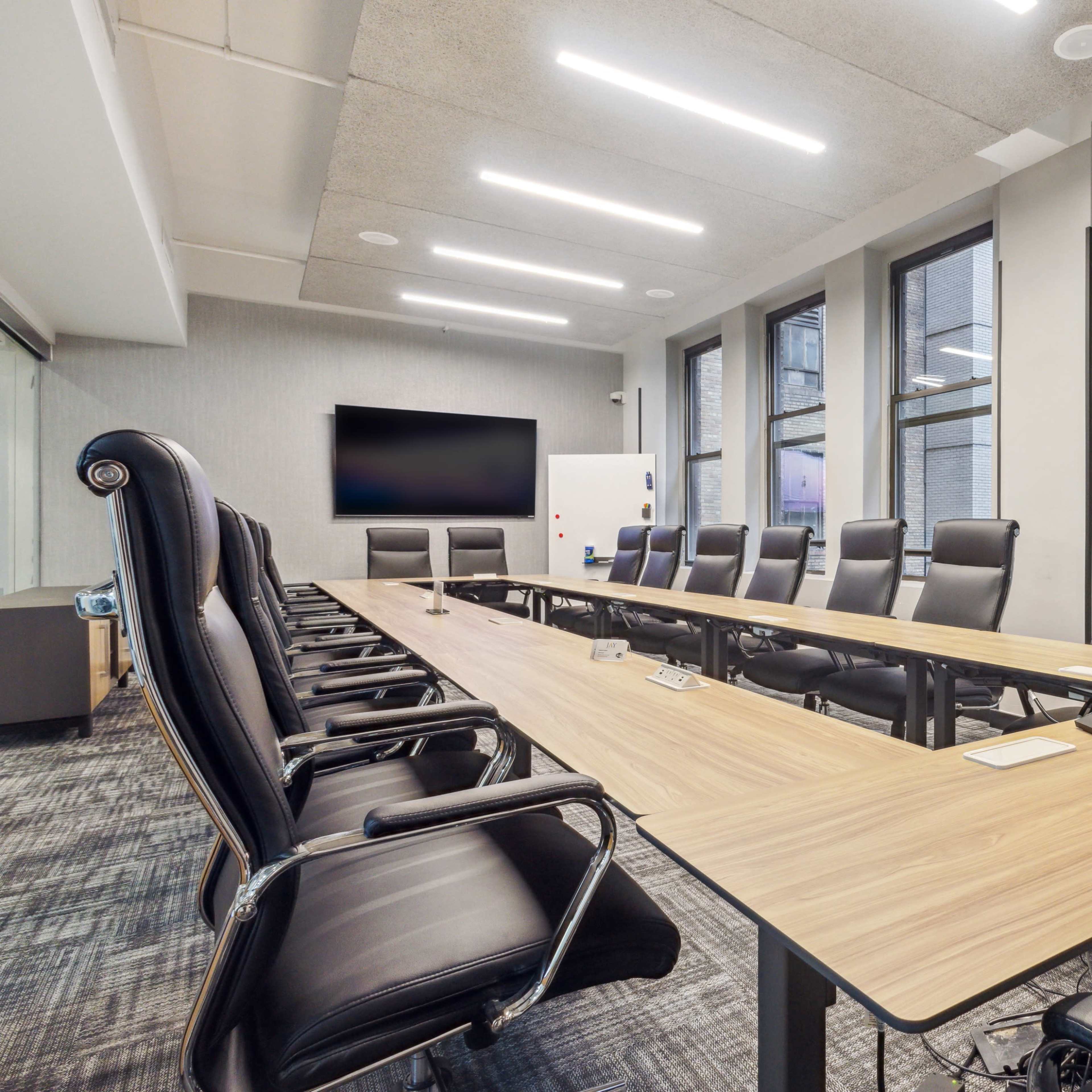A modern conference room features a long wooden table surrounded by black leather chairs and large windows providing natural light.