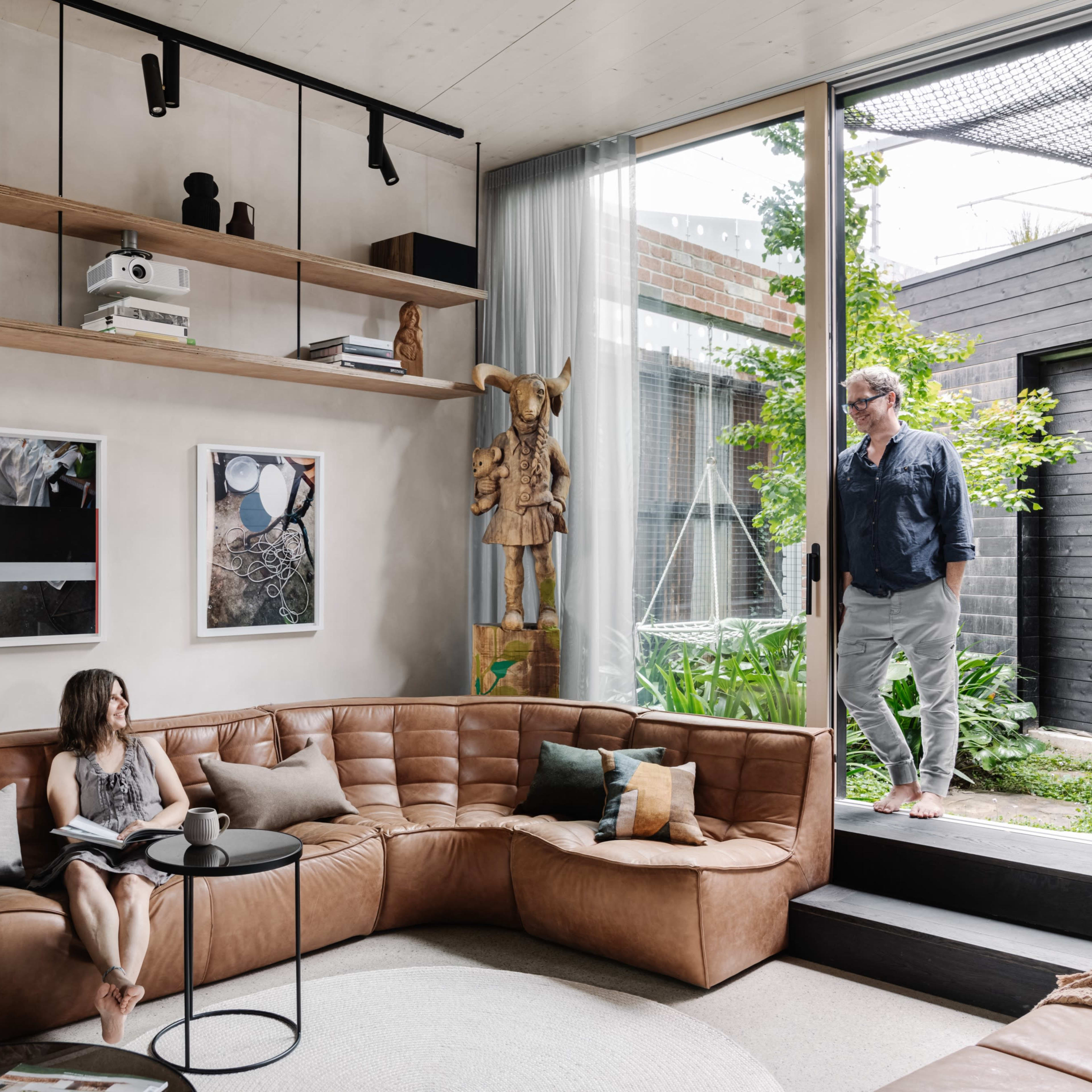 A man stands outside in a garden while a woman sits on a brown leather sectional sofa, reading a book in a modern living room.