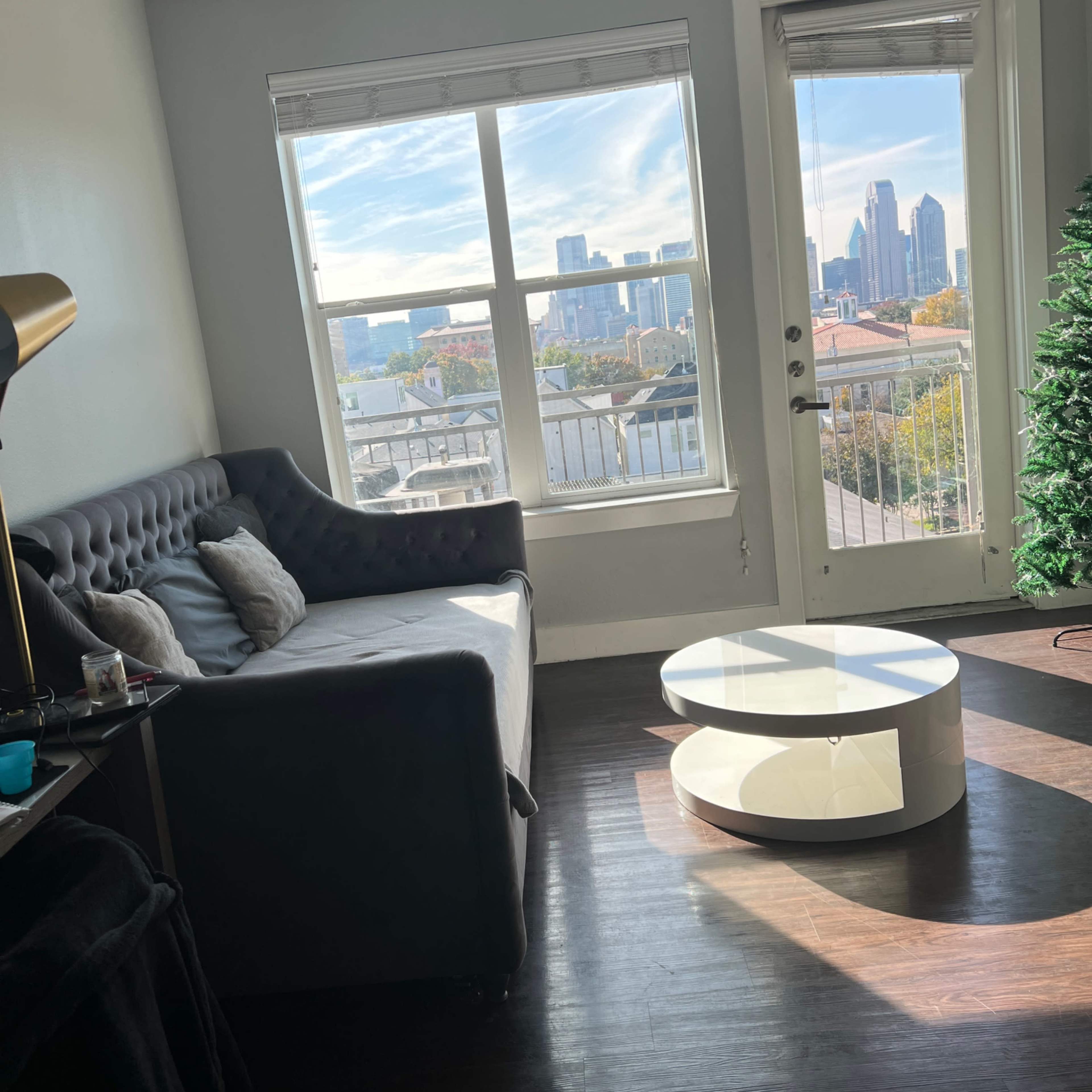 A living room with a gray couch, a circular white coffee table, and large windows providing a view of a city skyline.