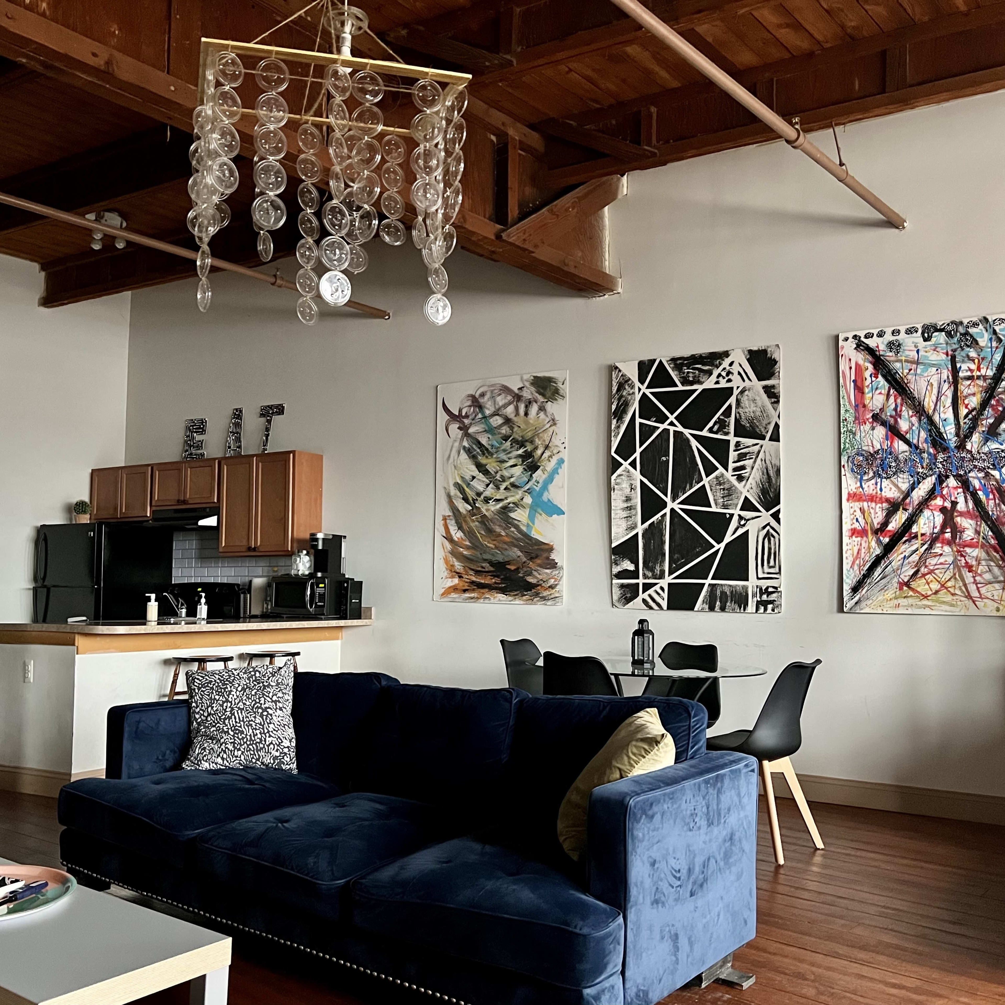 A spacious living area with a blue couch, a modern dining table and chairs, artistic wall hangings, and a kitchen area in the background.
