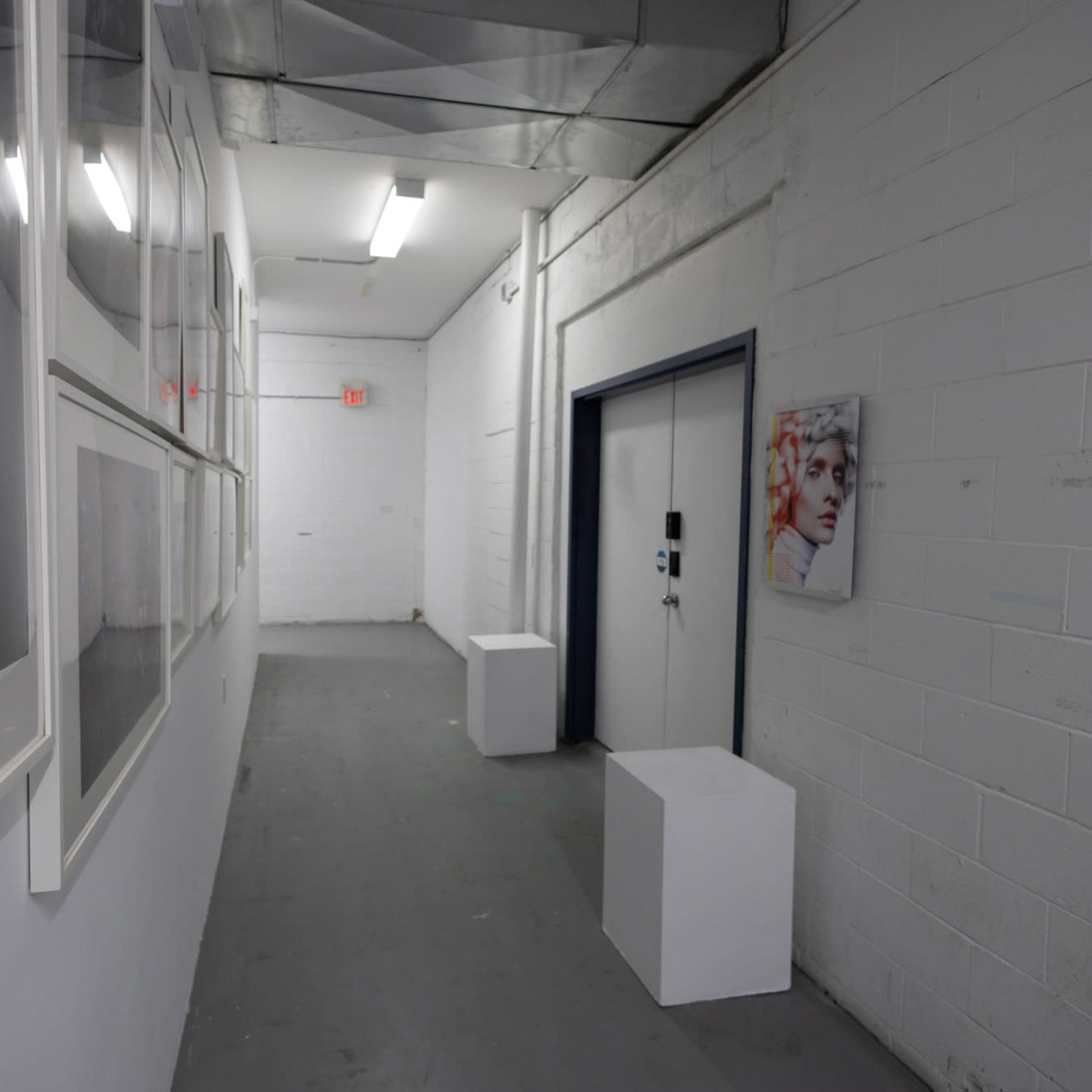 A narrow hallway with white walls features framed photographs on one side and a door with a small portrait hanging adjacent to it.