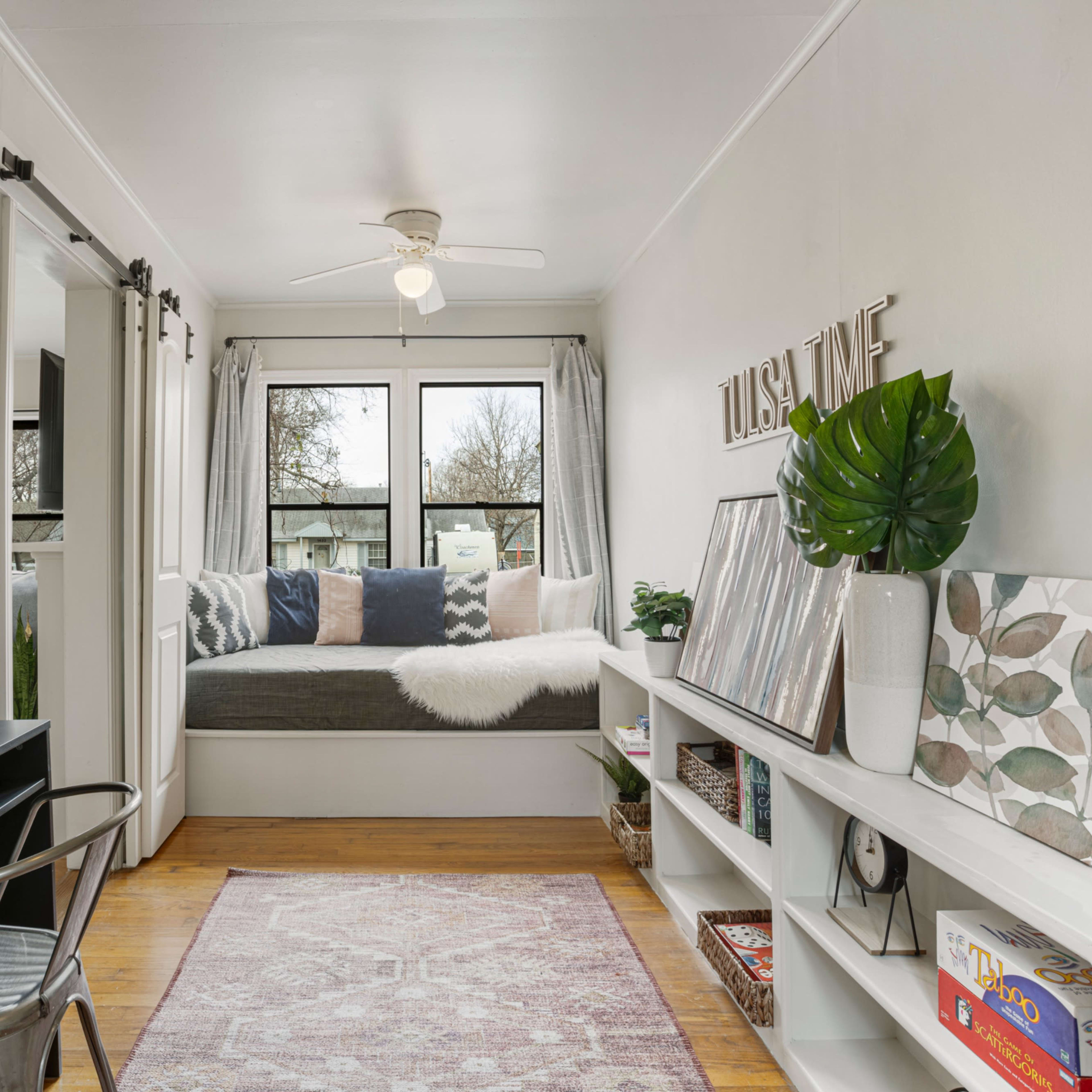 A cozy room features a window seat with cushions, a desk on the left, and a shelf displaying decorative items and plants.
