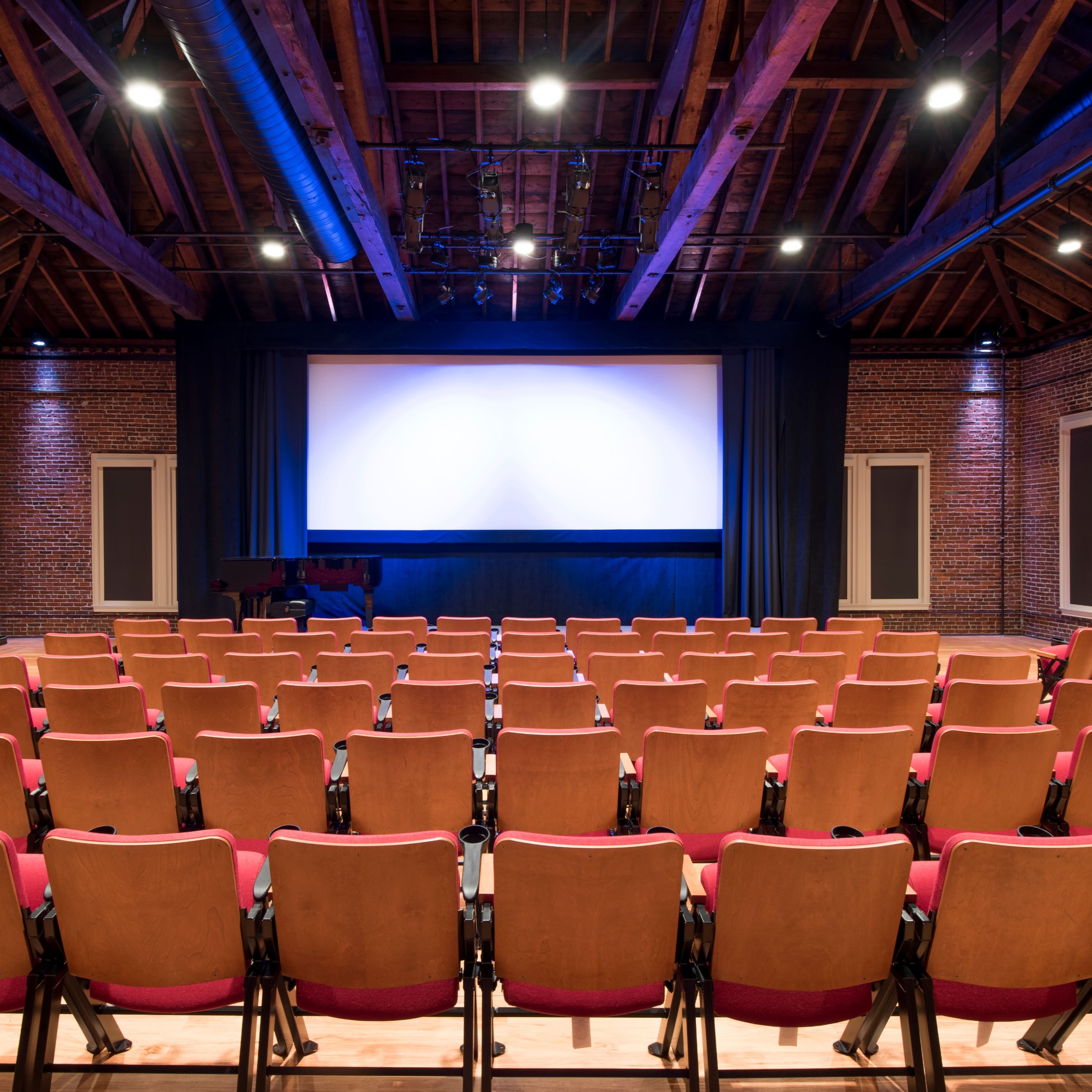 An empty auditorium with wooden beams, brick walls, and rows of seating facing a large screen.