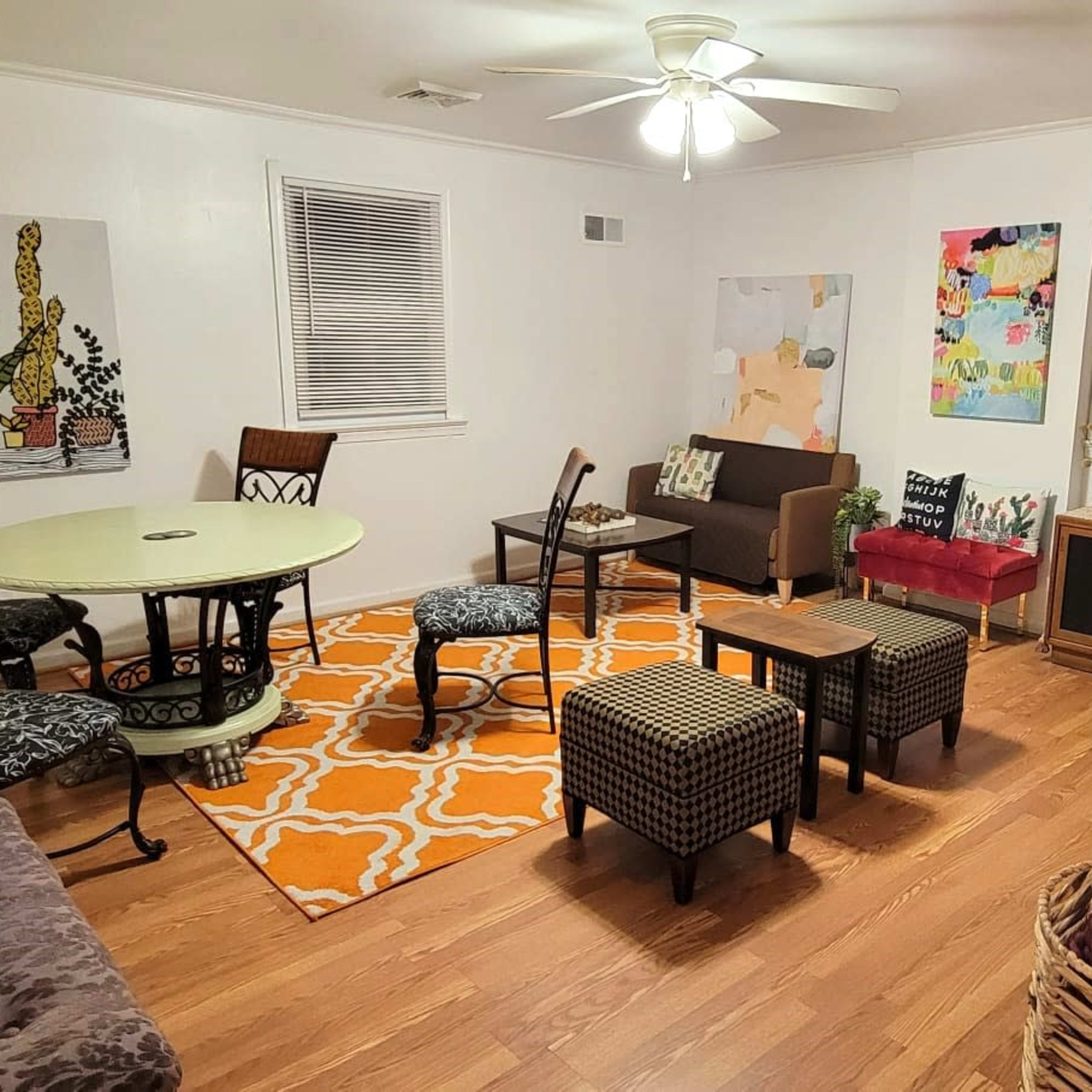 The room features a circular table surrounded by several chairs, with artwork on the walls and a mix of seating arrangements on a patterned rug.