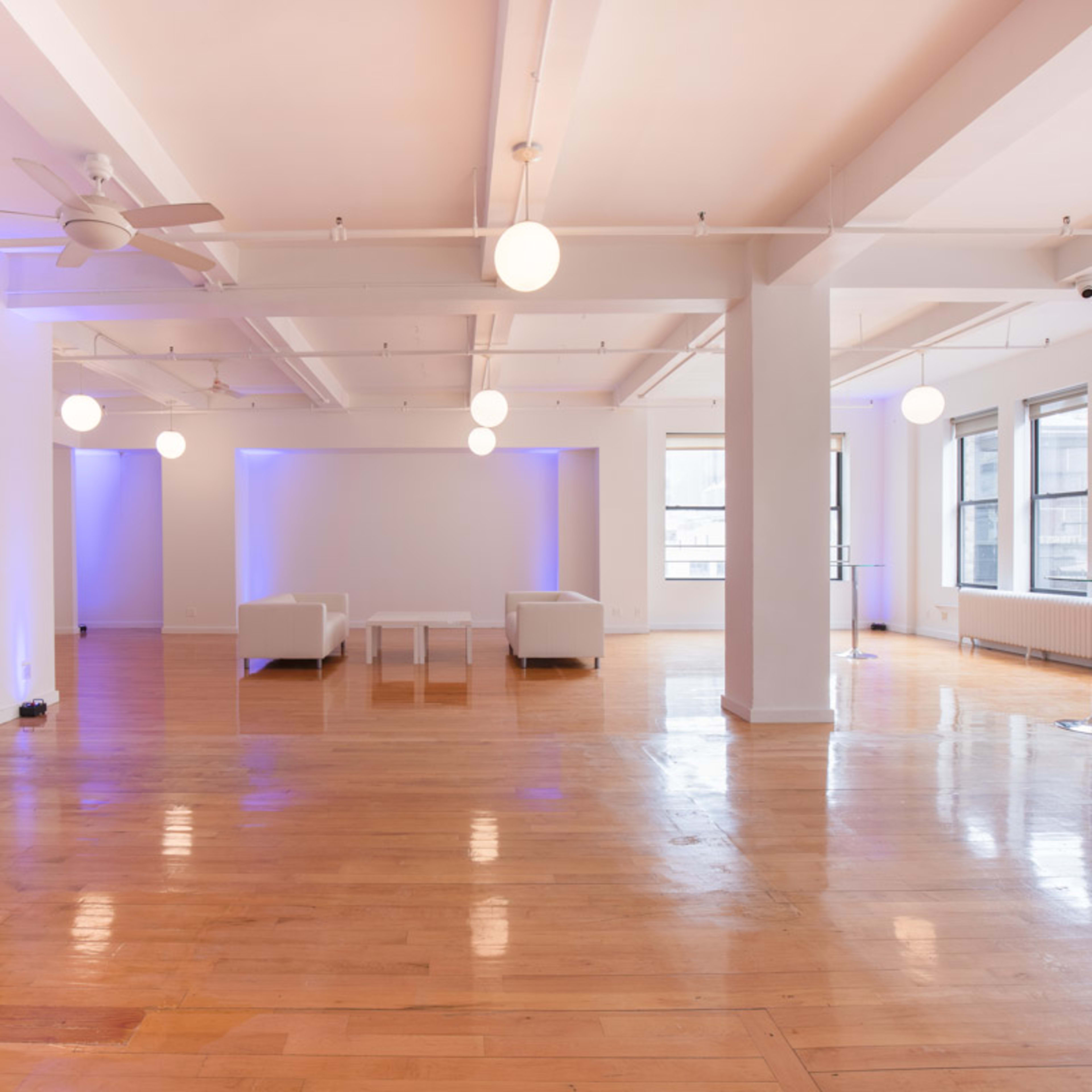 A spacious, well-lit room features wooden floors, white walls, and minimalist furniture, with several round light fixtures hanging from the ceiling.