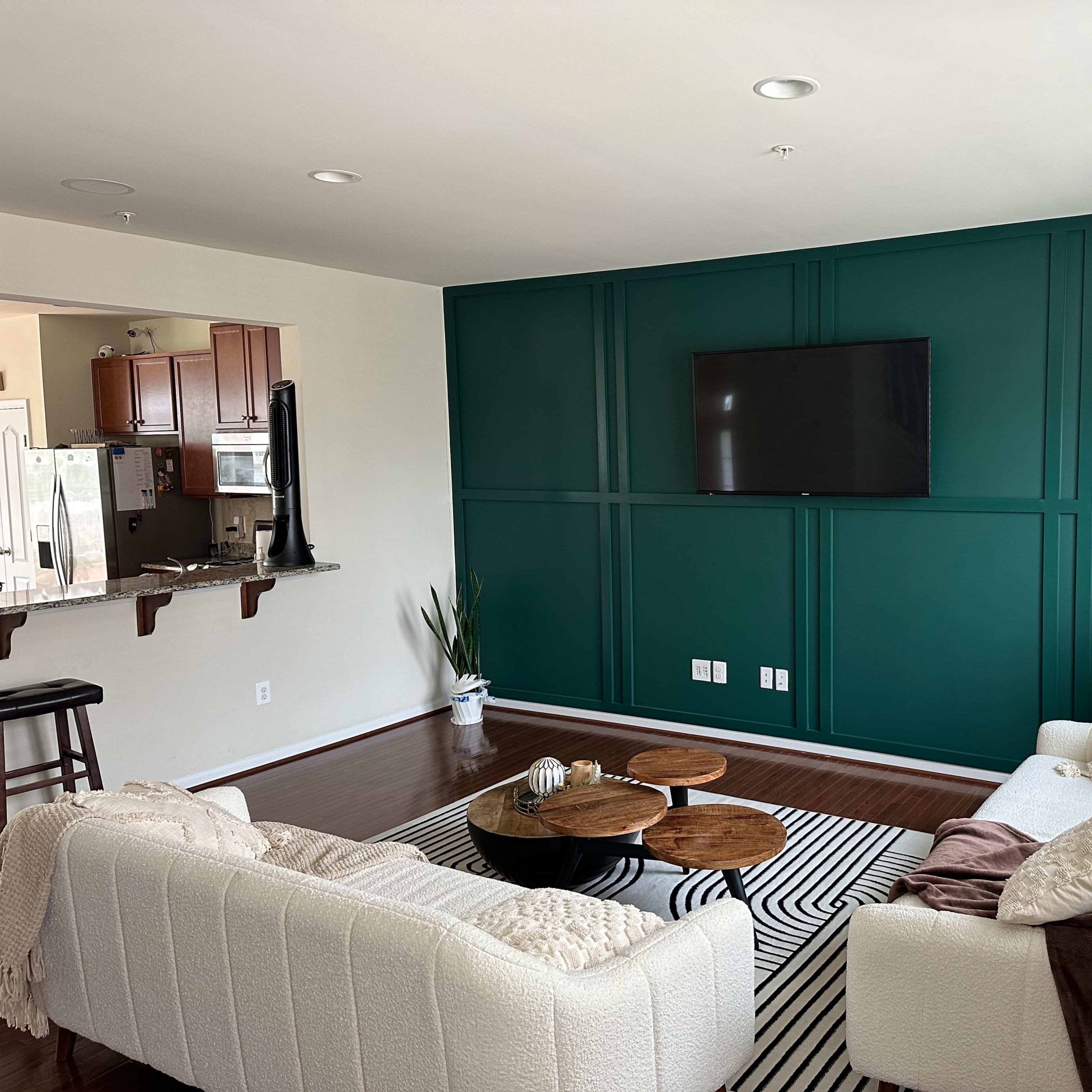 A modern living room with a dark green accent wall, a television mounted on it, a light-colored sofa set, and a striped area rug on the wooden floor.