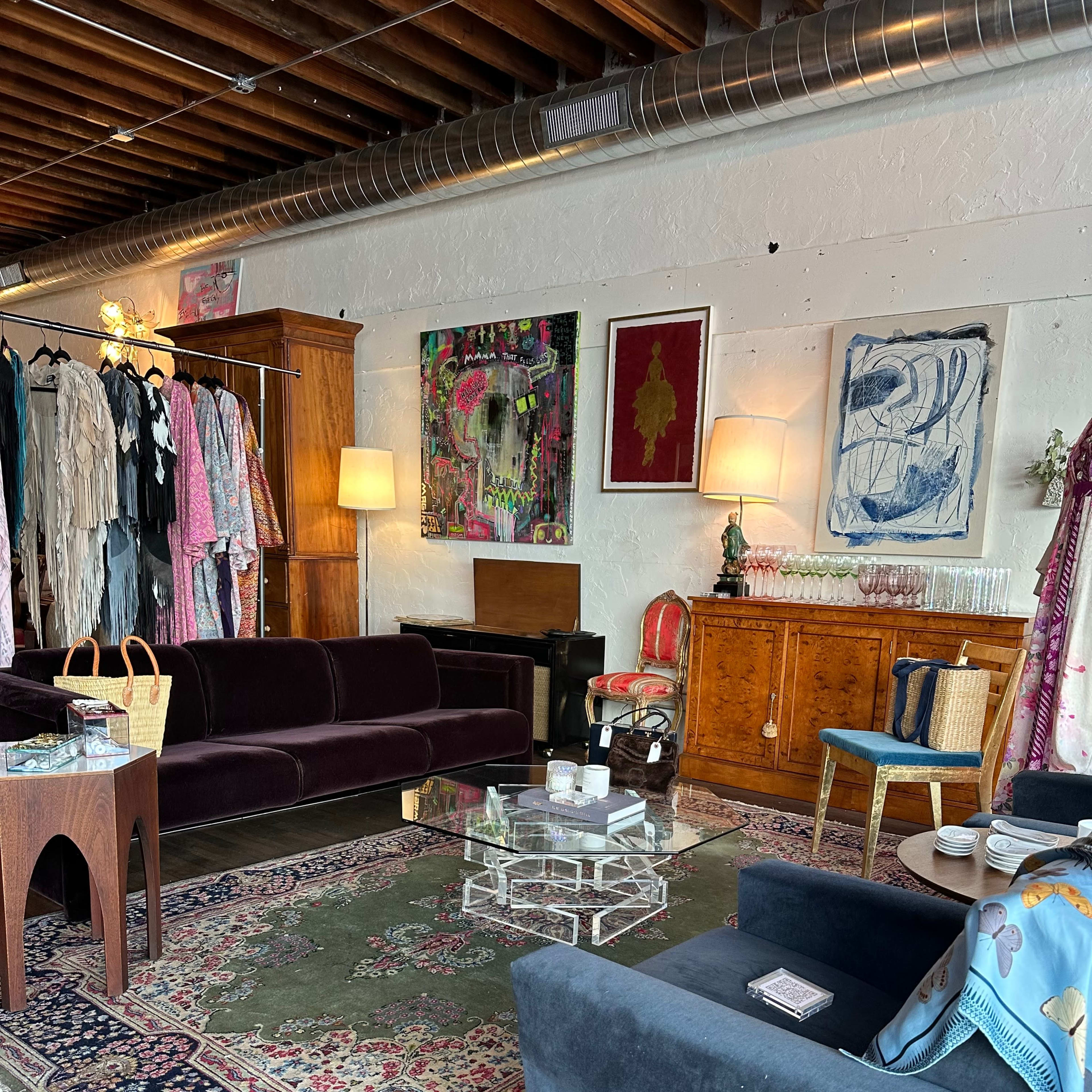 A stylish boutique interior featuring a mix of clothing racks, artwork on the walls, a plush sofa, and a glass coffee table set on a patterned rug.