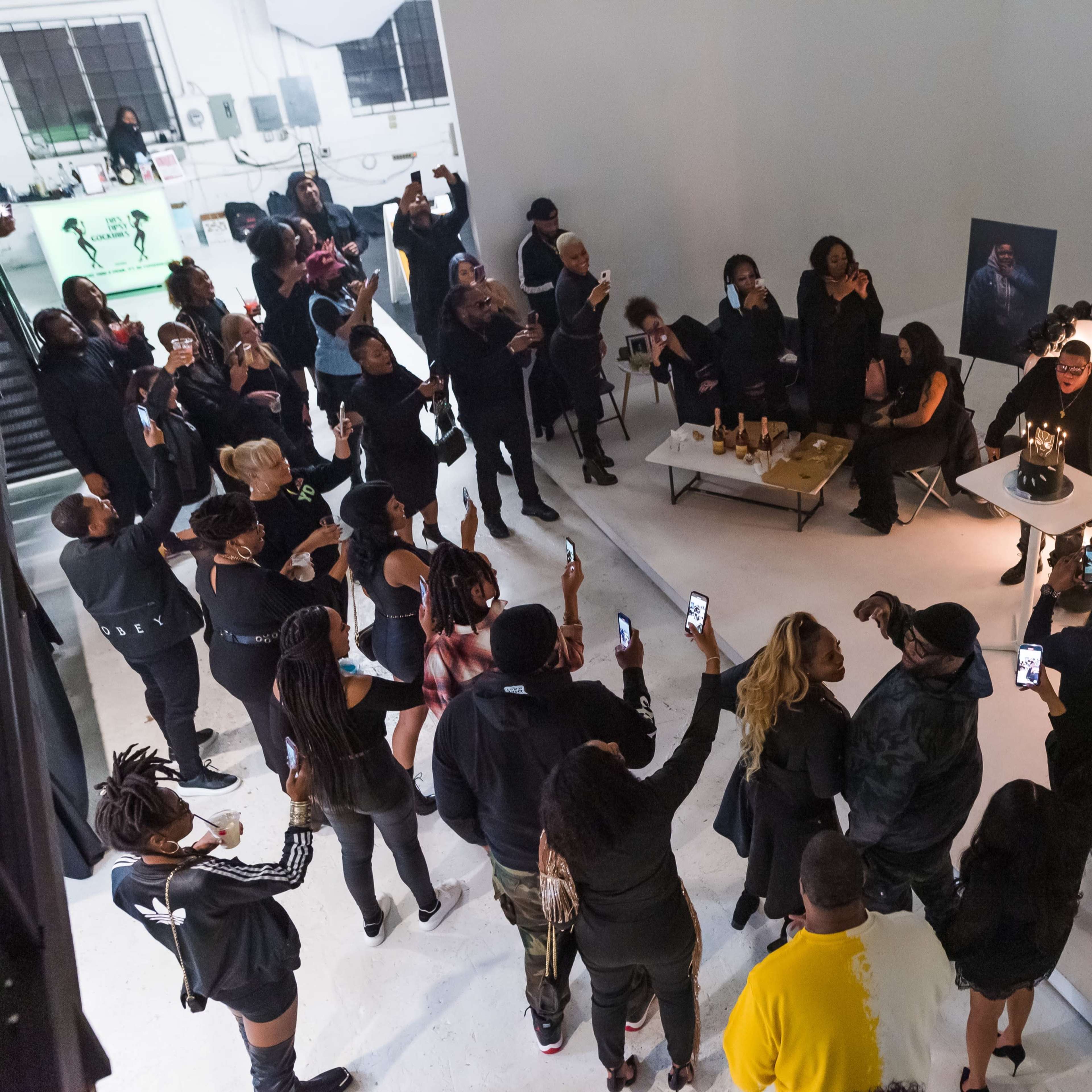 A crowd of people is gathered in a gallery celebrating a birthday milestone, with a cake and decorations prominently displayed.
