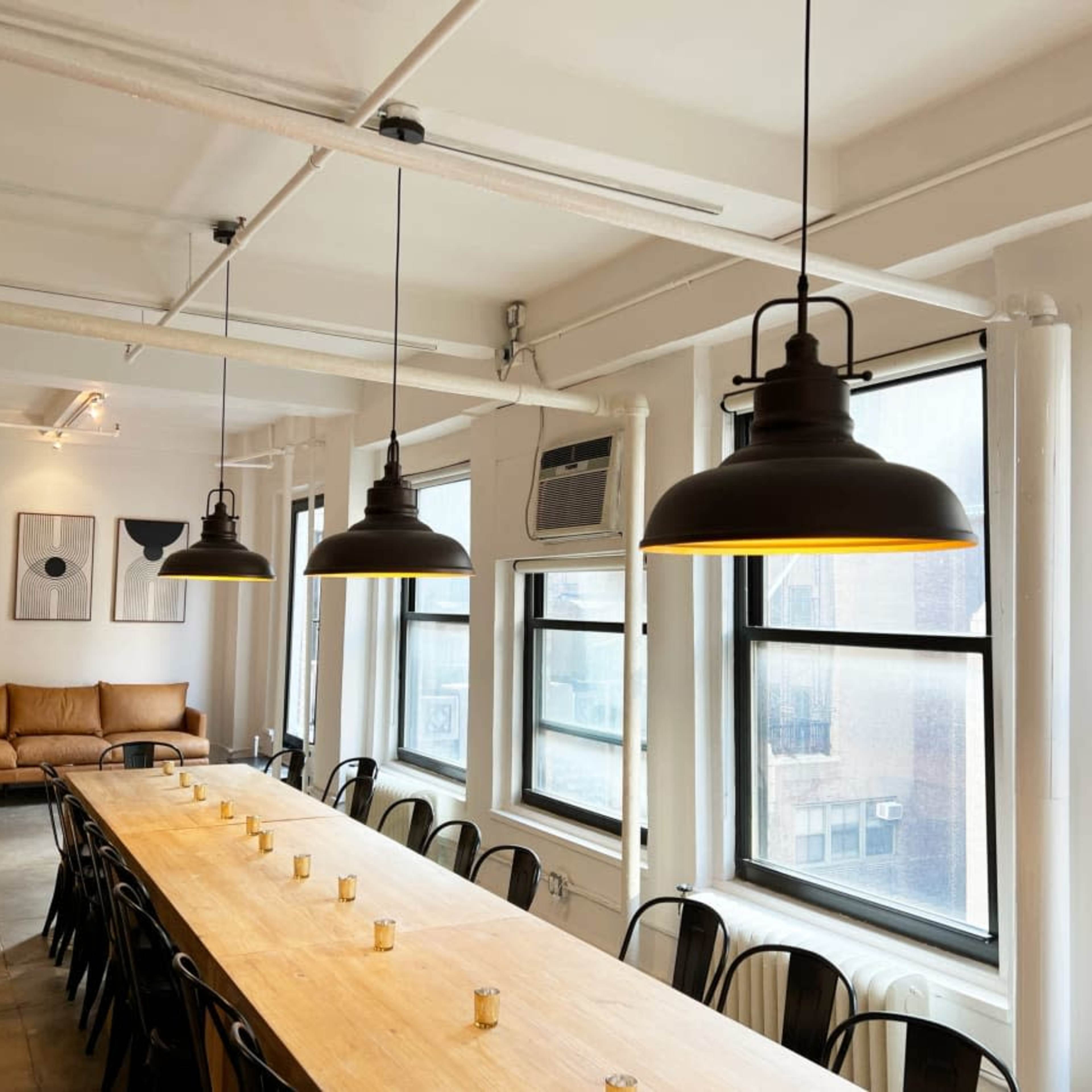 A spacious meeting room featuring a long wooden table, black chairs, three hanging pendant lights, and a Christmas tree in the corner.