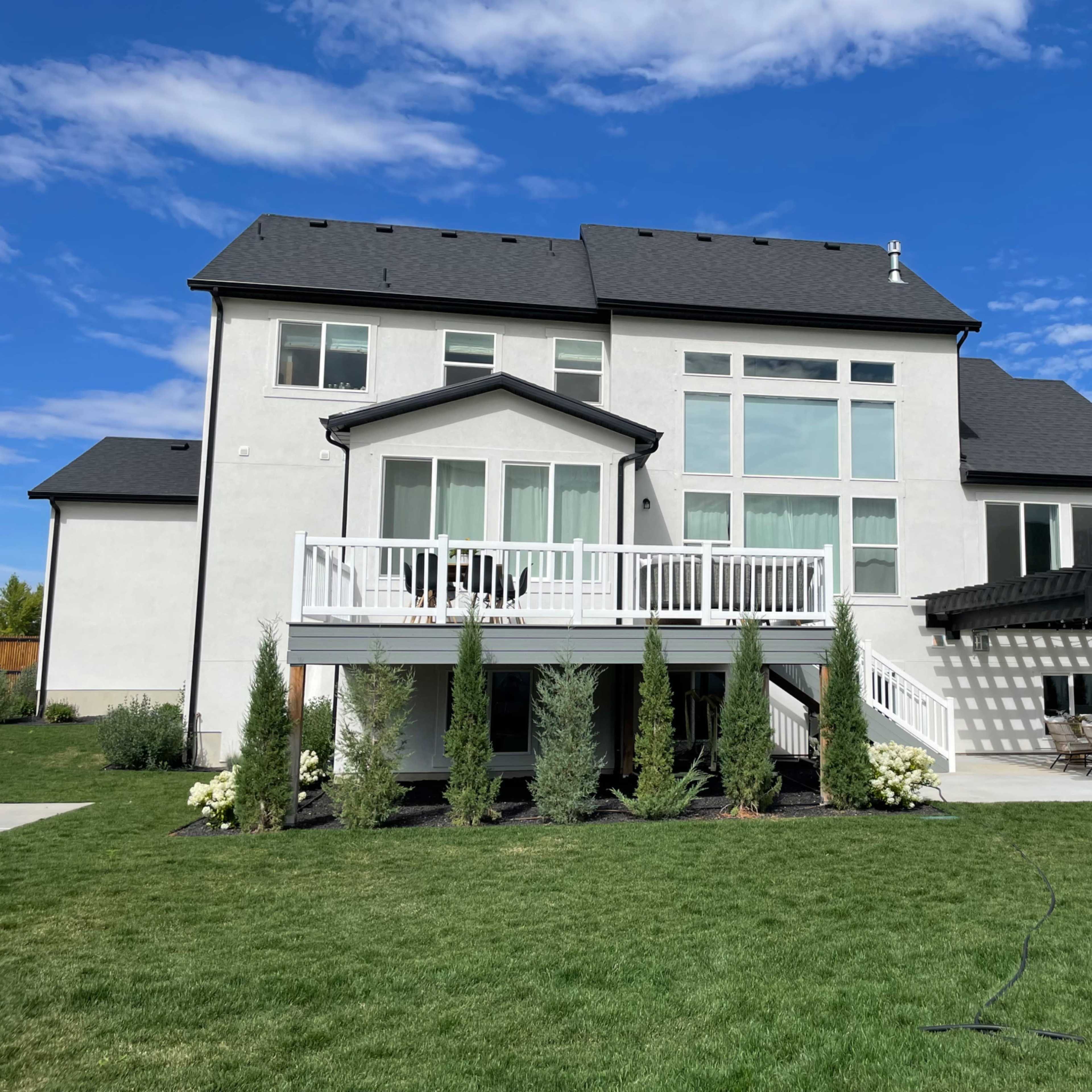A large two-story house with a white exterior, multiple windows, and a deck overlooks a neatly landscaped yard.