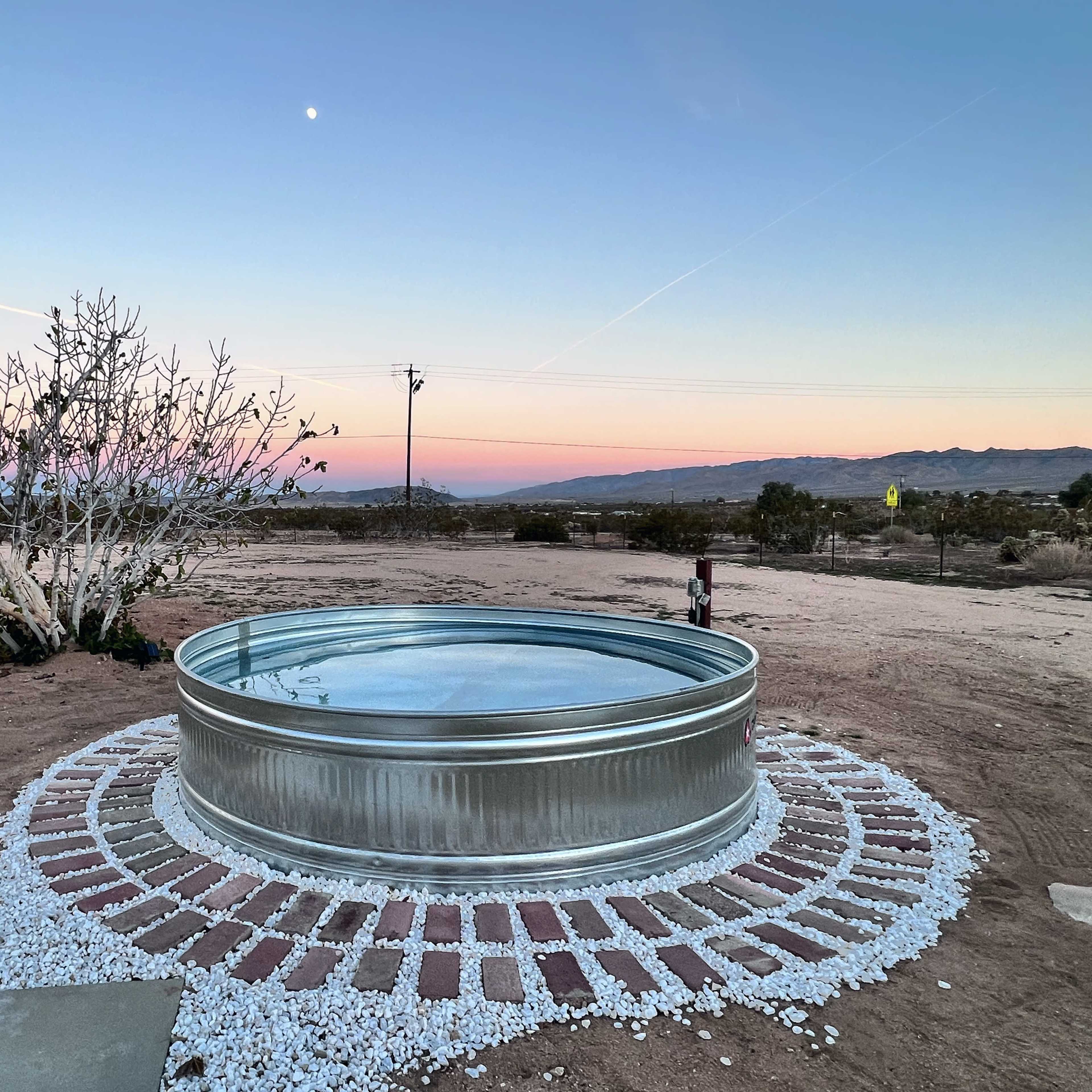 A circular metal water tank surrounded by a stone and gravel border sits in a barren landscape with a clear sky at dusk.
