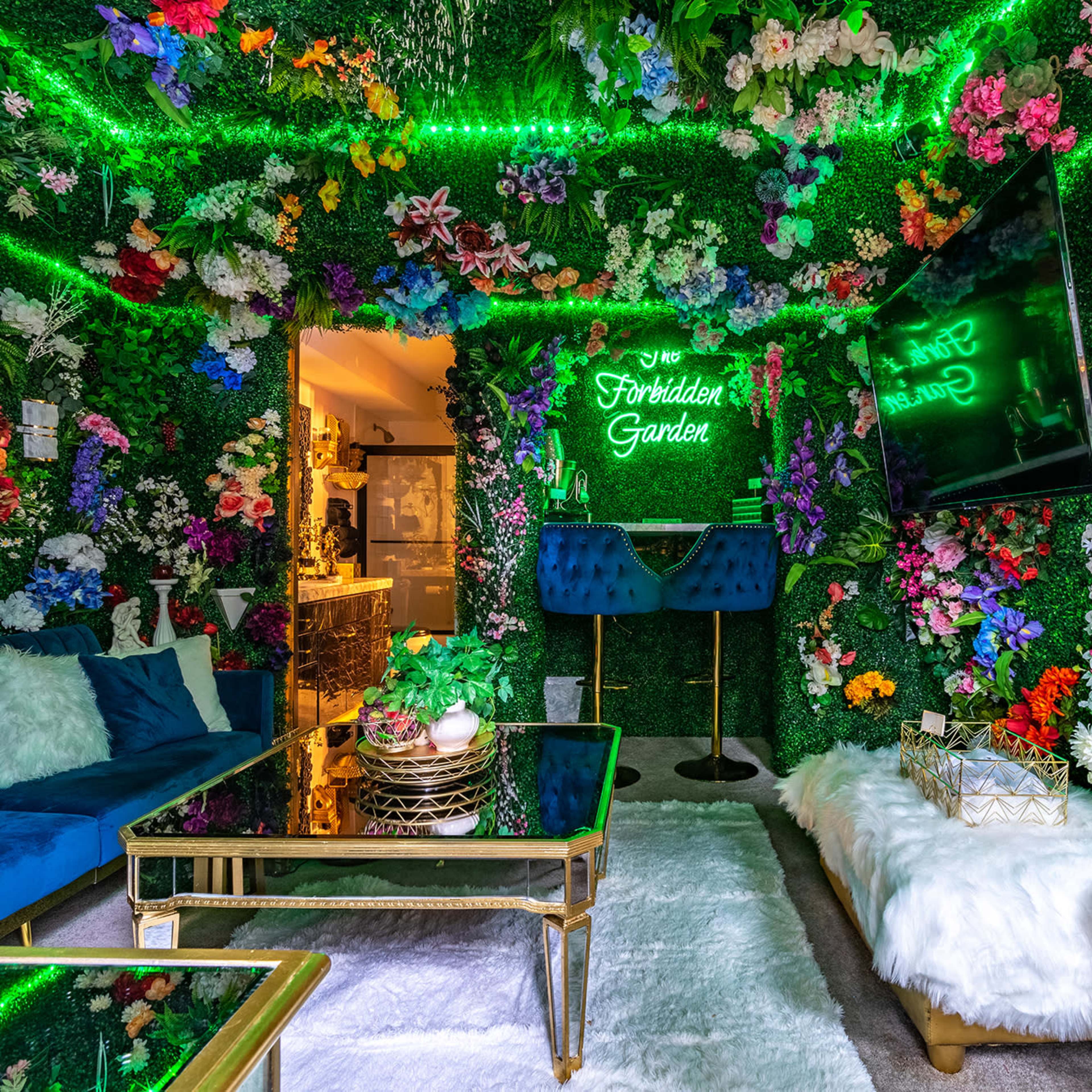 The room features walls covered in vibrant artificial flowers, a plush white rug, a glass coffee table, and two blue velvet chairs, with a neon sign reading "Forbidden Garden."