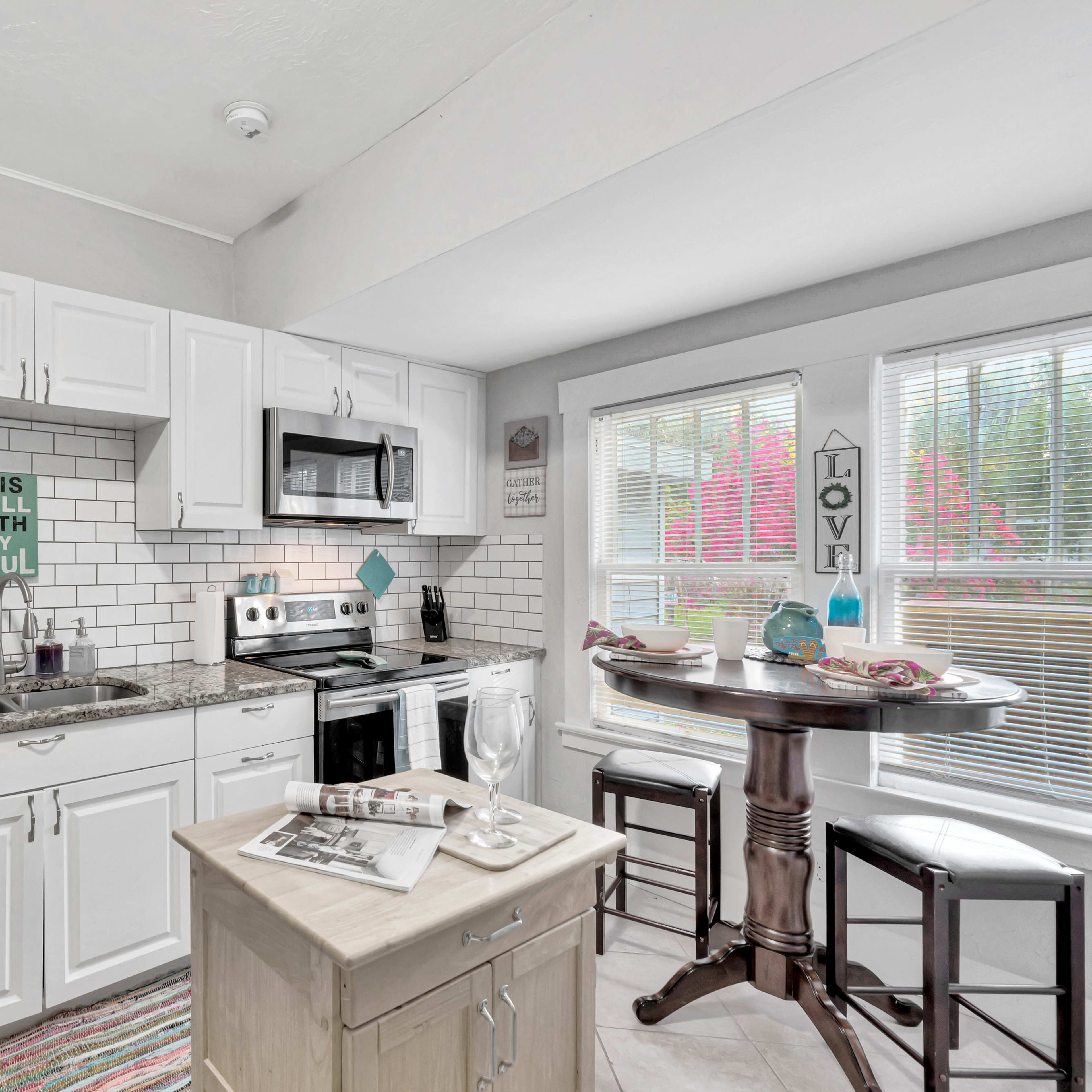A modern kitchen with white cabinets, a gray countertop, a small dining table by the window, and tile backsplash.