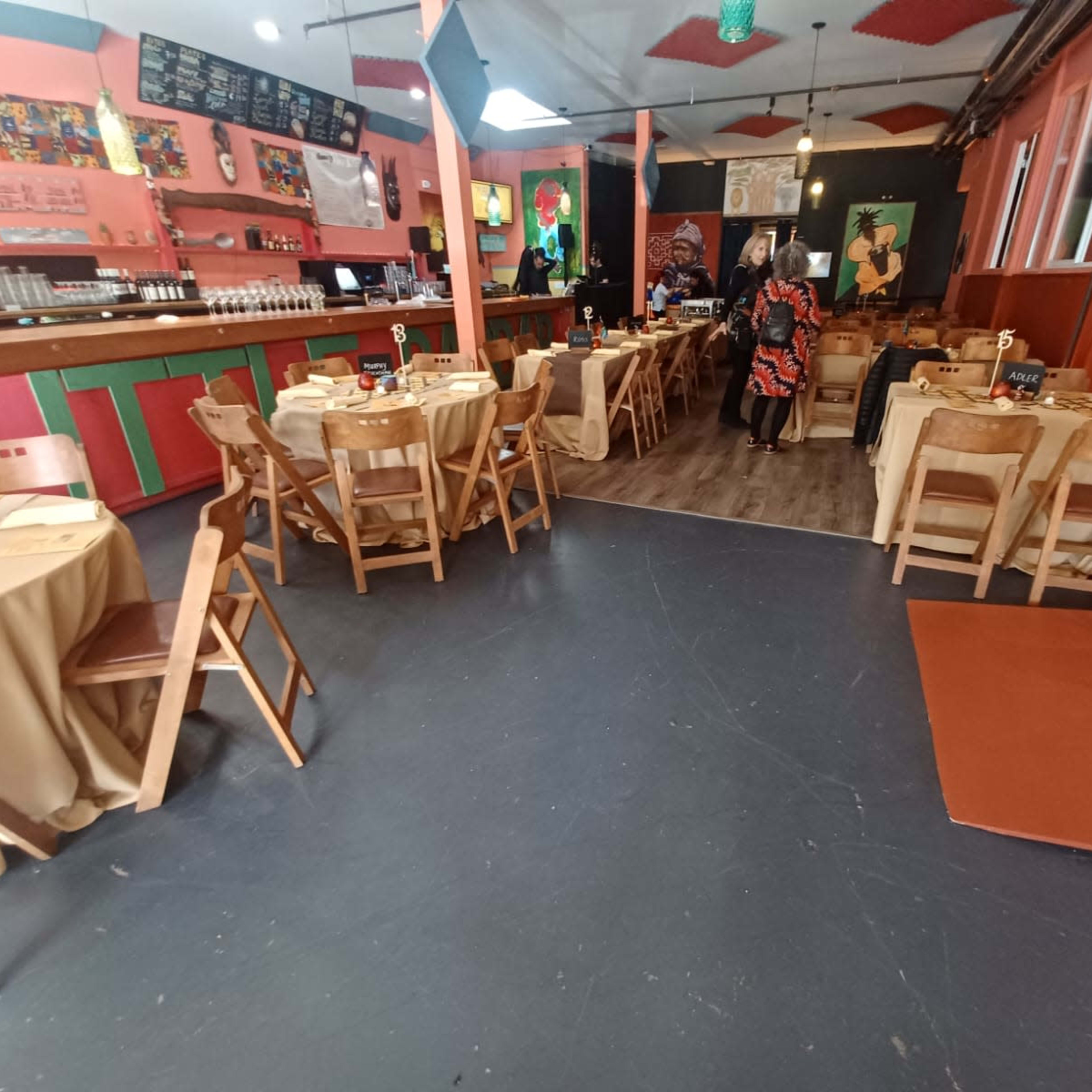 The interior of a restaurant with wooden tables and chairs set up, featuring a brightly colored decor and a bar area in the background.