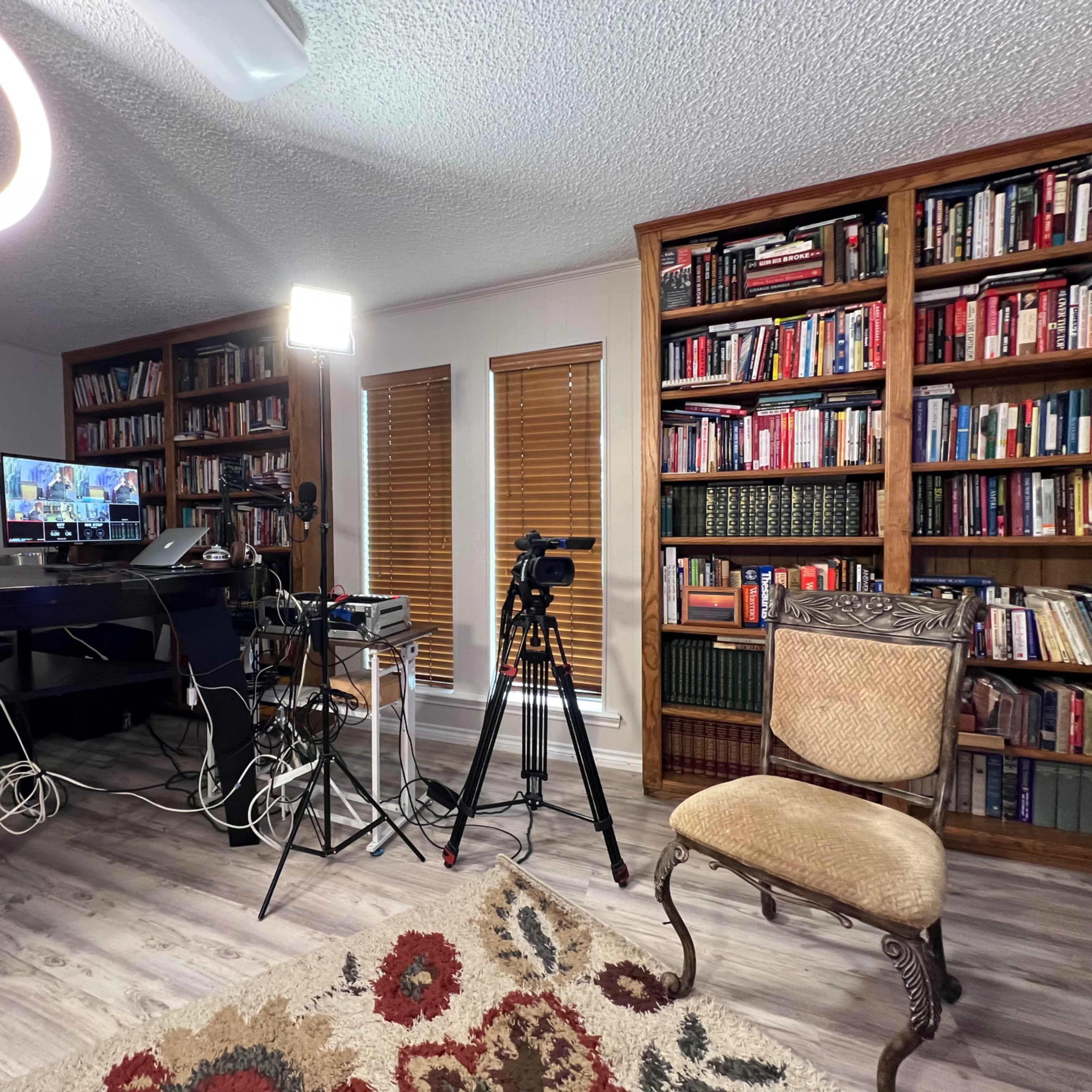 A room with a desk, multiple cameras, and light equipment set up next to a bookshelf filled with books, with an empty chair positioned nearby.