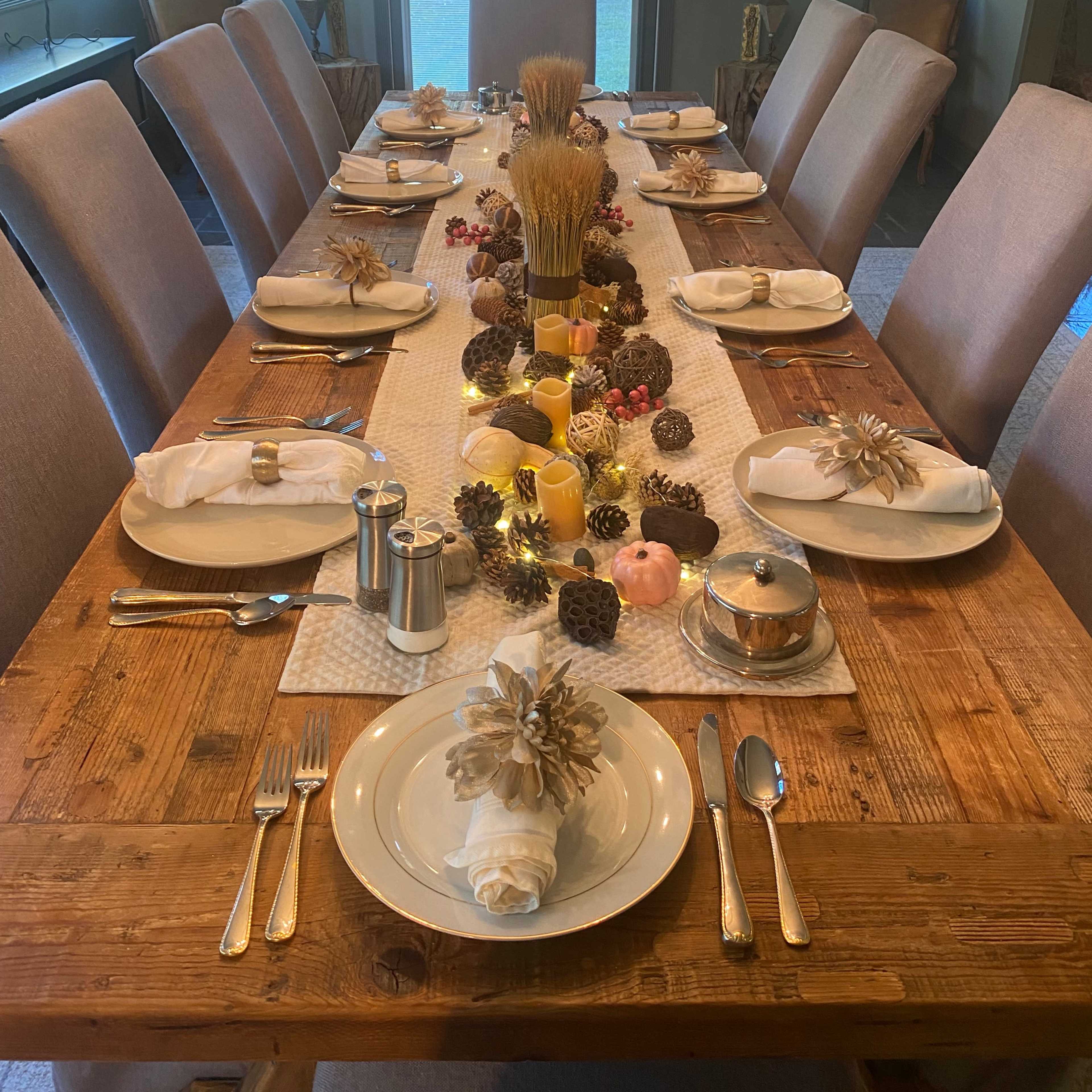 A long wooden dining table is elegantly set with white plates, silverware, and a decorative centerpiece of pine cones, candles, and seasonal decorations.