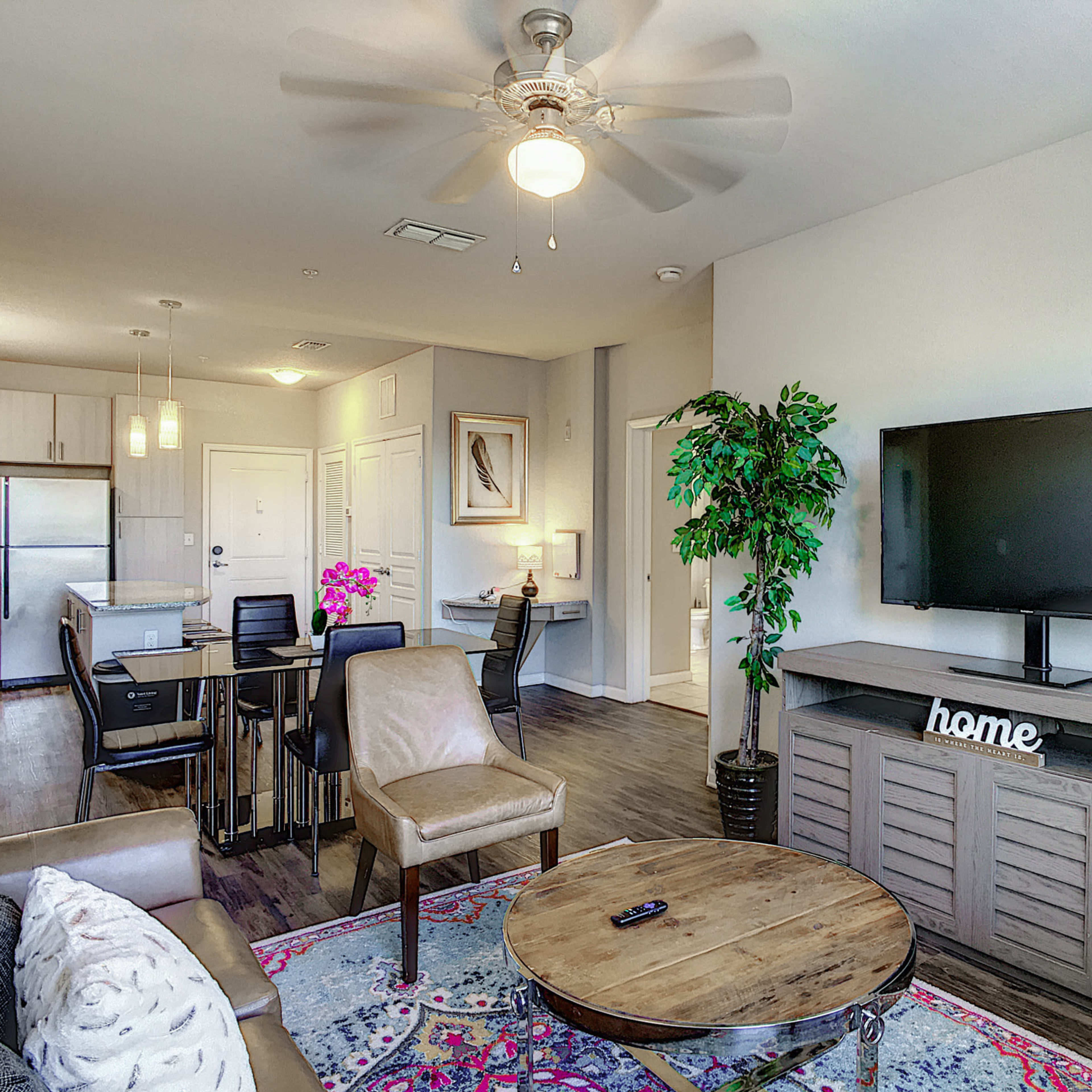 A modern living room and dining area featuring a comfortable seating arrangement, a round coffee table, a large television on a media console, and a kitchen visible in the background.