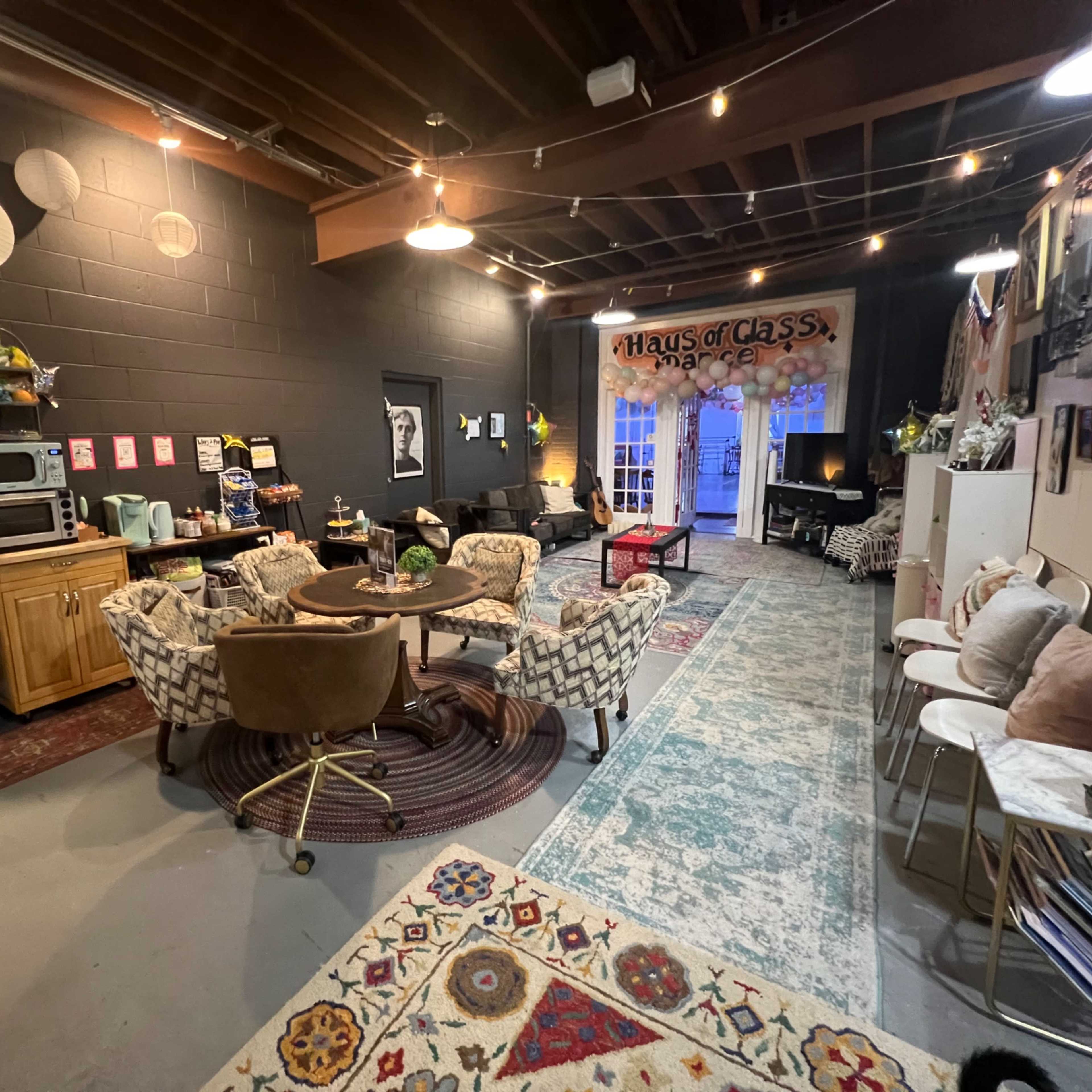 A cozy interior space with a mix of seating arrangements, decorative rugs, and bright lighting, featuring a kitchen area and artwork on the walls.