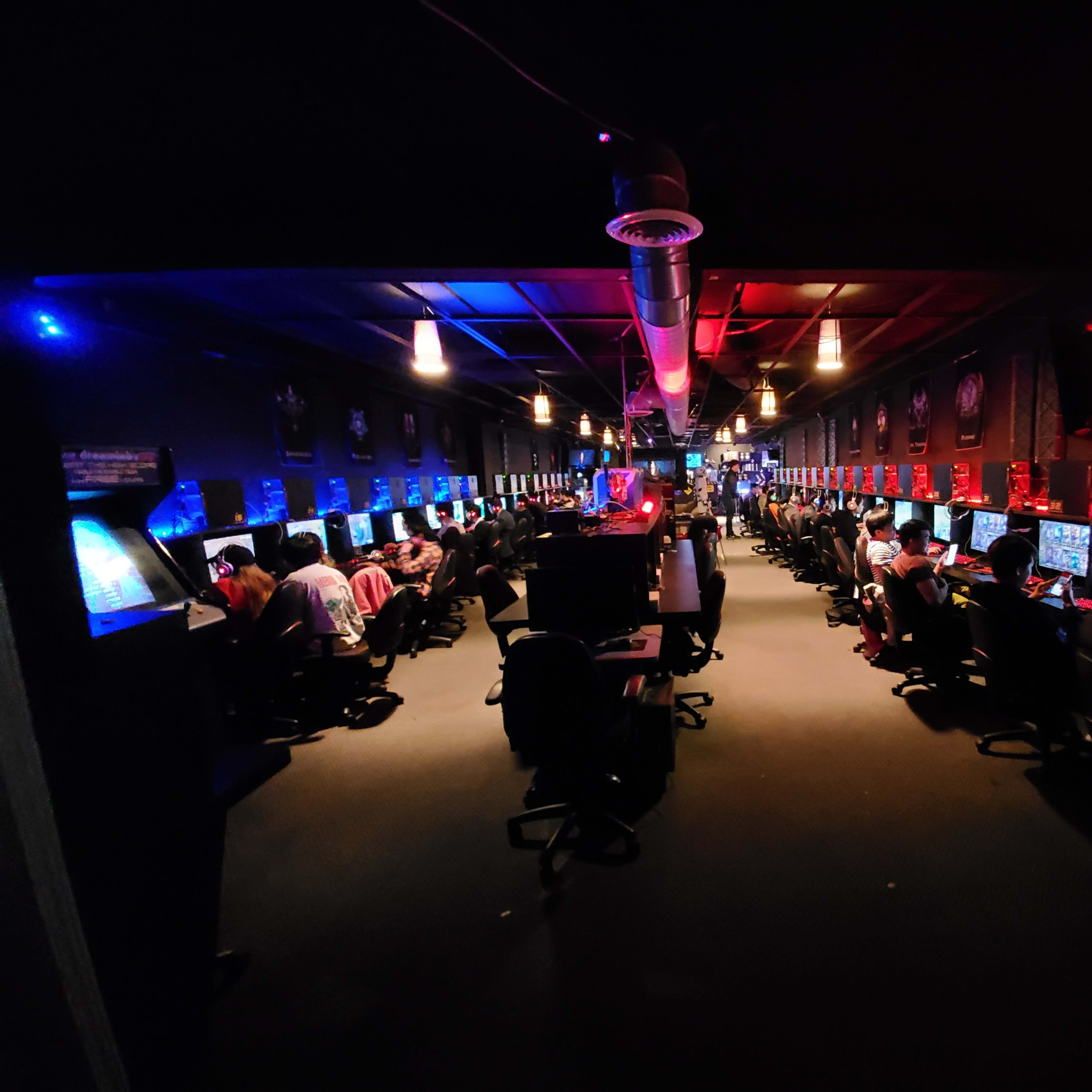 A crowded gaming café with multiple gaming stations, each equipped with computers, set in a dimly lit room illuminated by colorful lights.