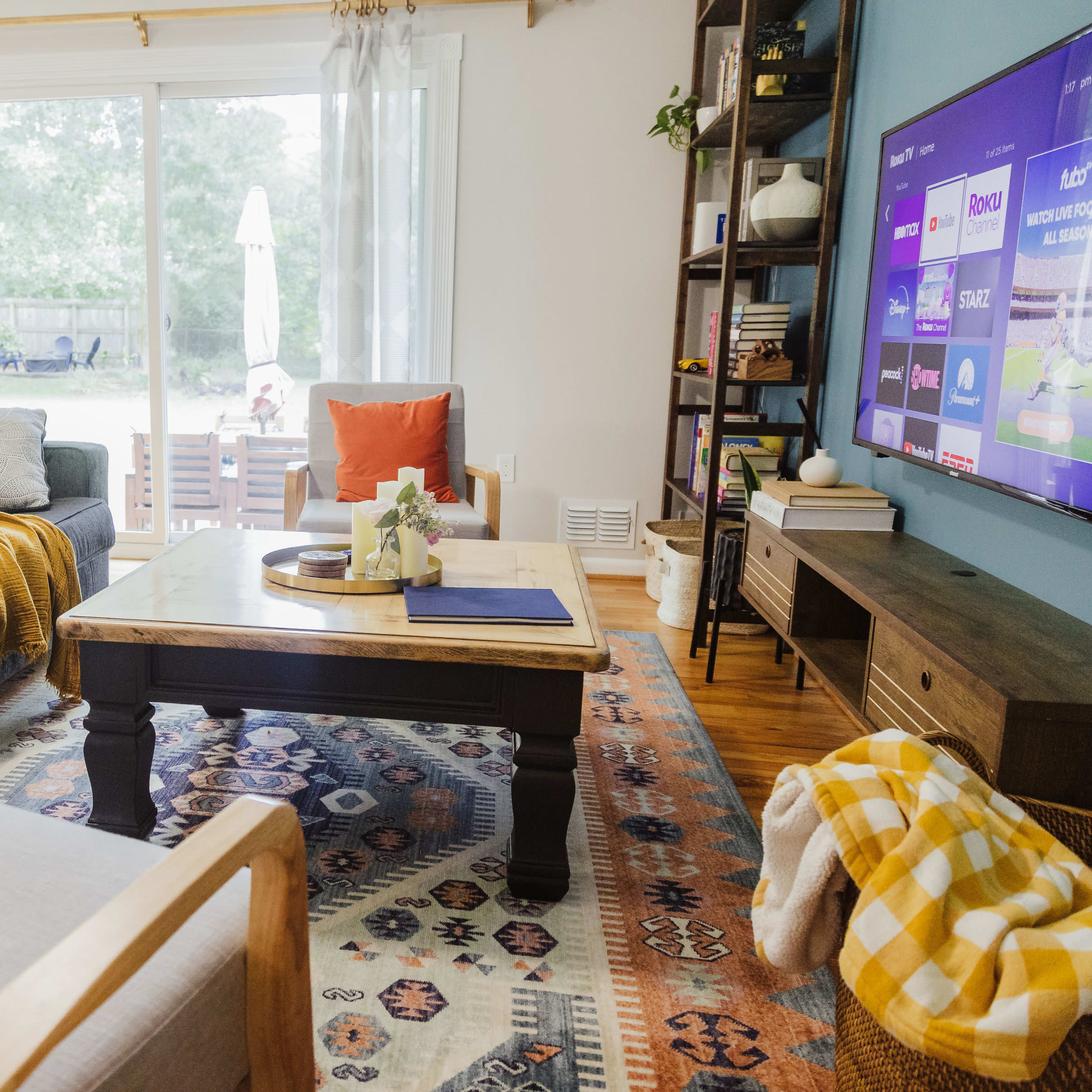 A cozy living room features a dark sofa with colorful throw pillows, a wooden coffee table on an area rug, and a large TV against the wall, with a view of an outdoor space through sliding glass doors.