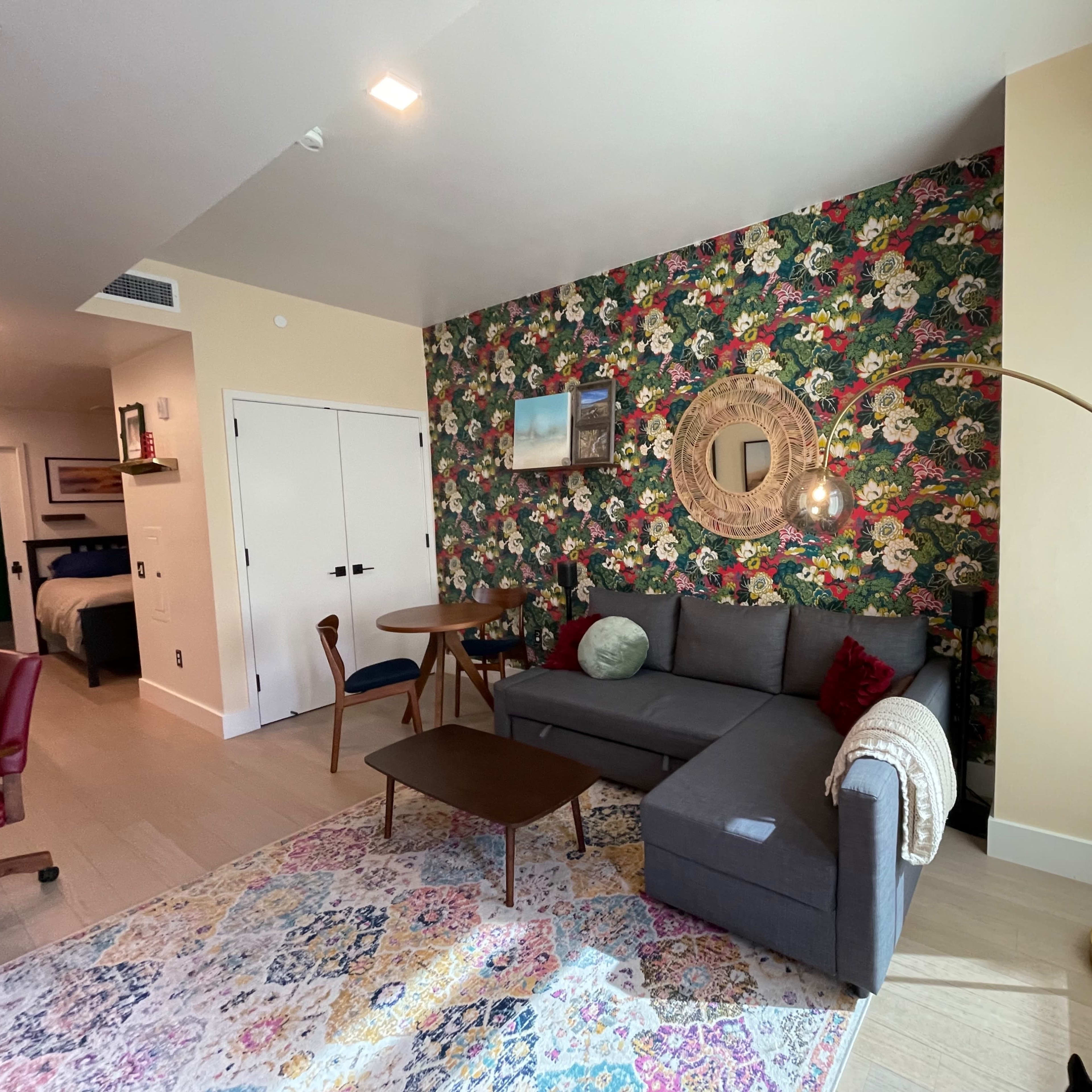 A living room featuring a floral accent wall, a gray sectional sofa, a round mirror, and a floor lamp beside a small table and chairs.