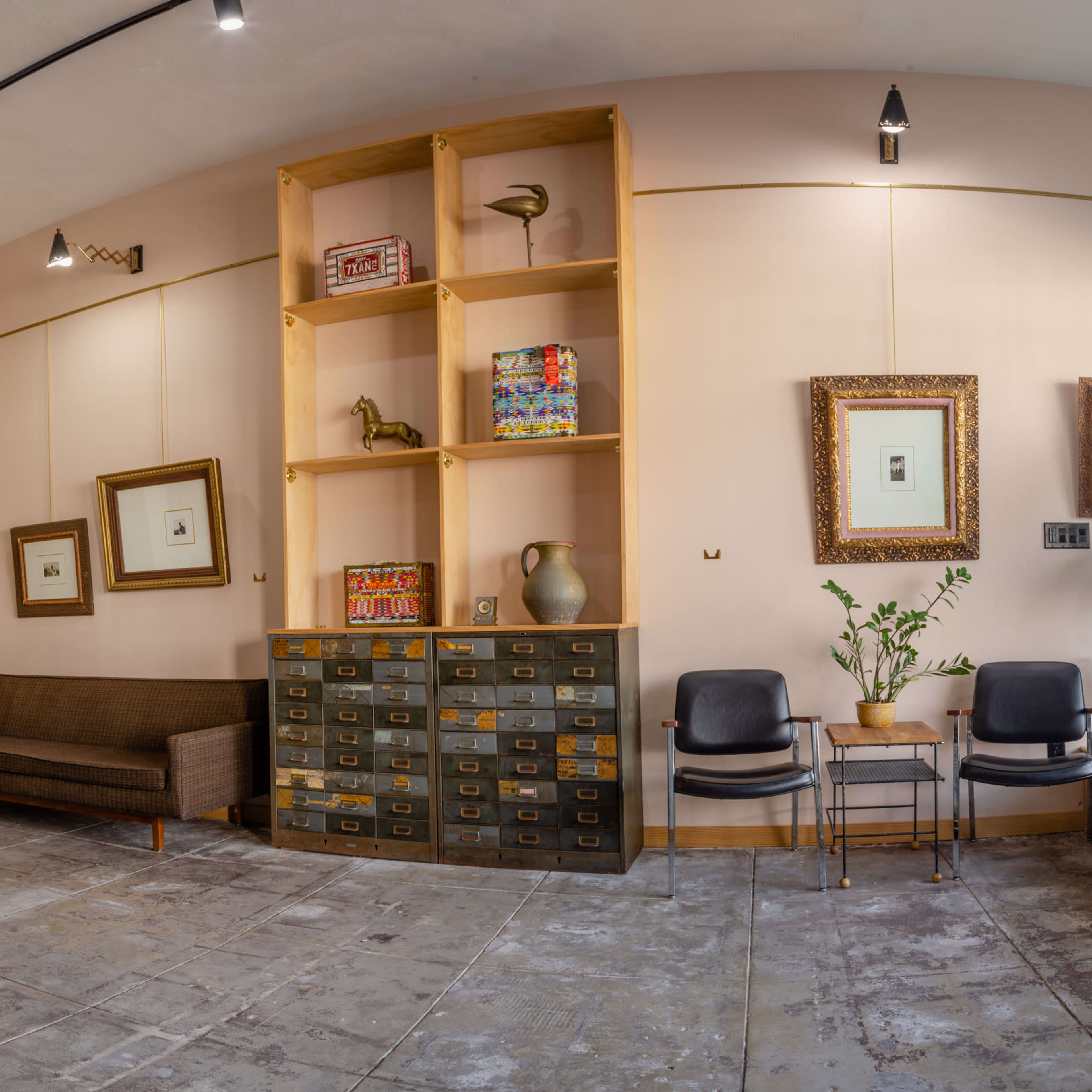 A modern interior space with a brown sofa, a tall shelving unit displaying items, framed artwork on the walls, and two black chairs near a potted plant.