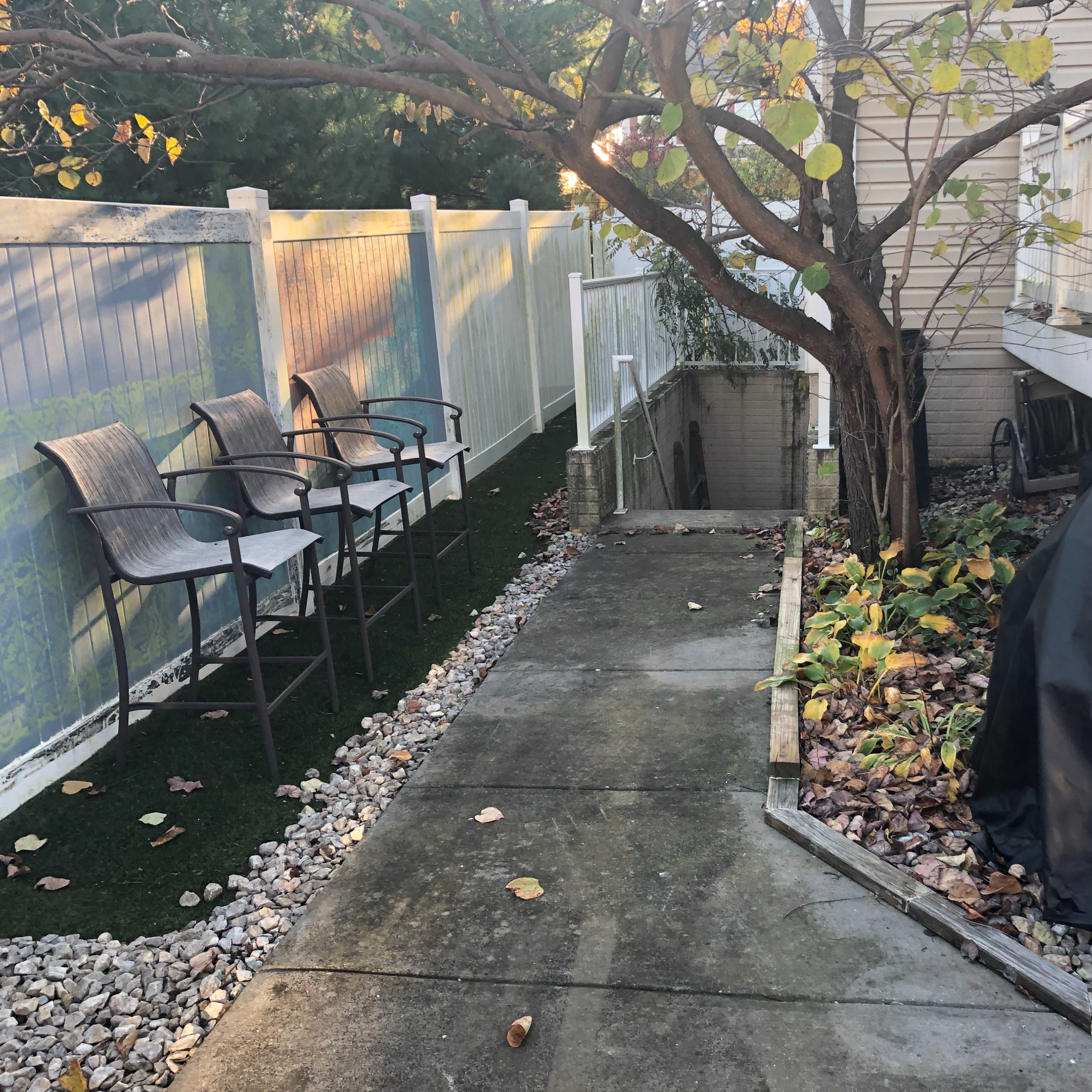 A concrete pathway runs alongside a fenced yard, flanked by chairs and a covered grill, leading to a garden area.