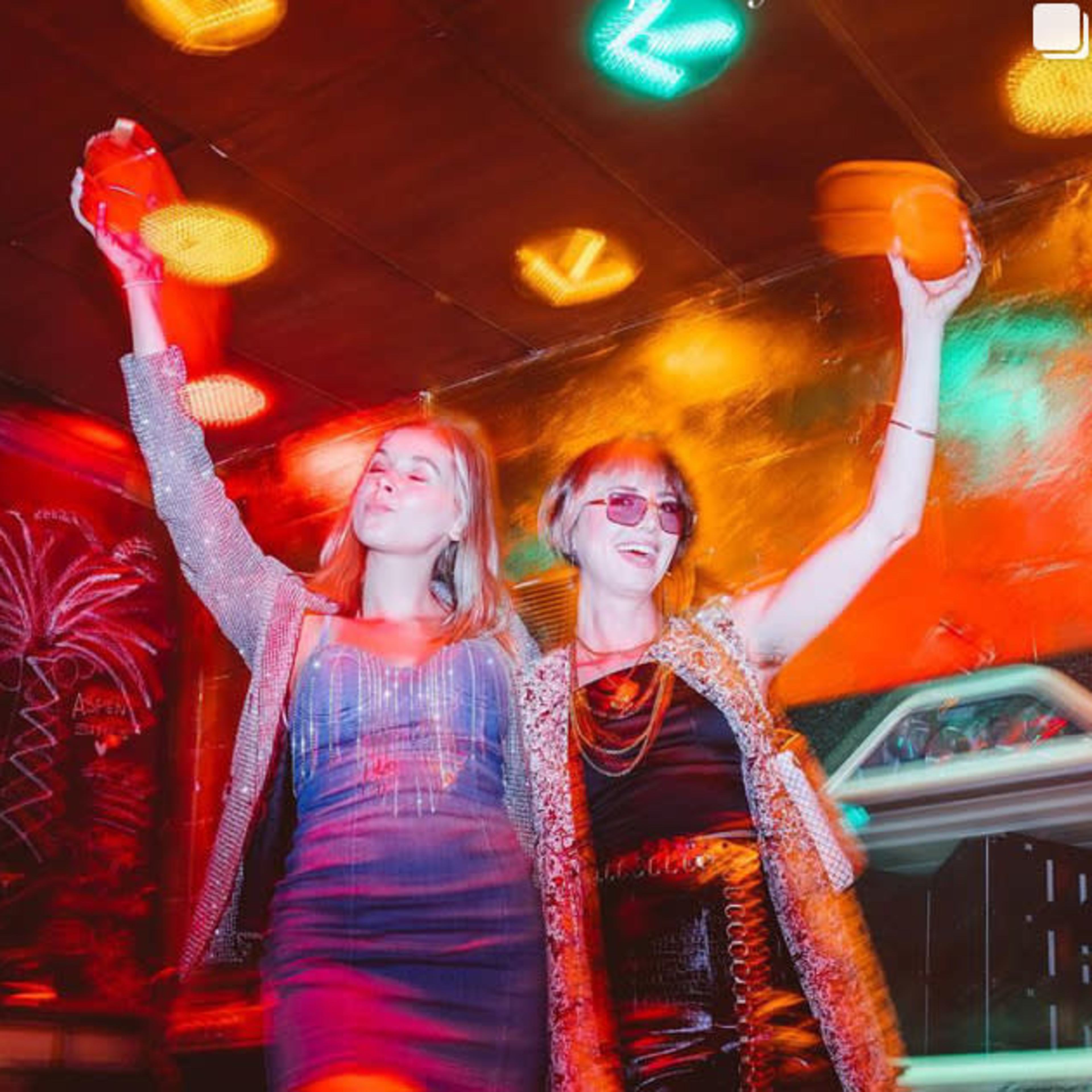 Two women are celebrating in a vibrant nightclub, holding drinks and wearing sequined outfits.