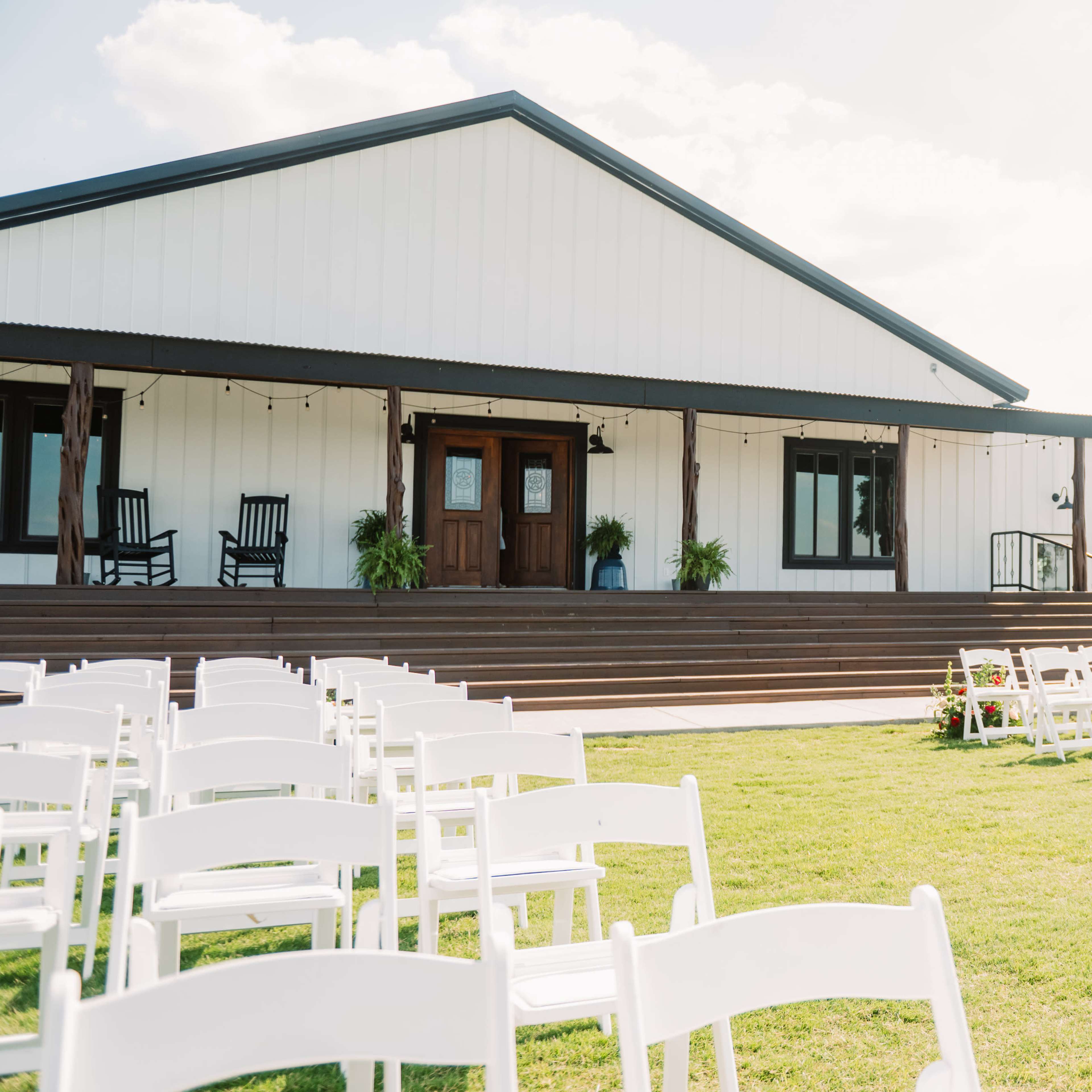 A white outdoor venue with a large building and rows of white chairs set up on a green lawn for an event.