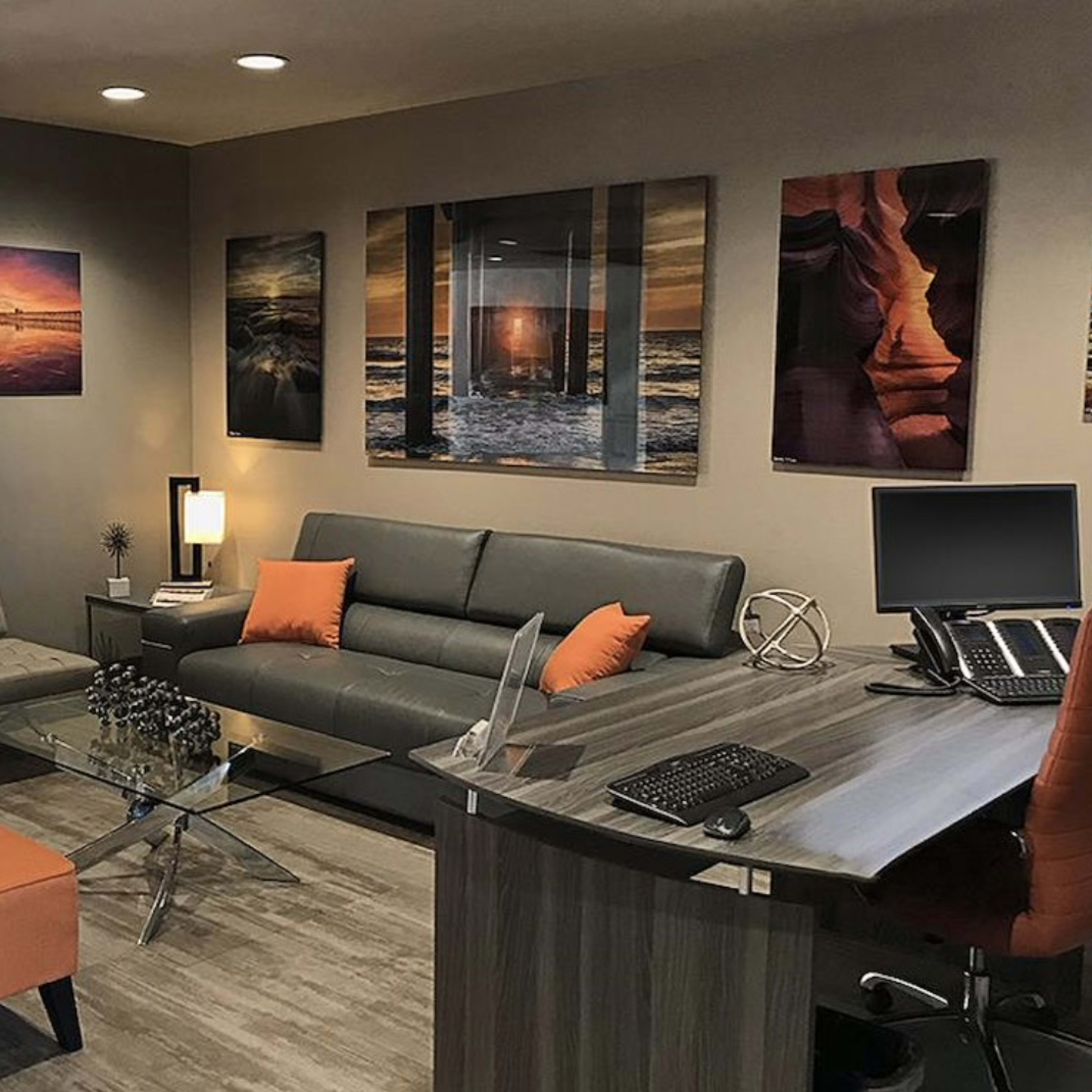 A modern waiting area featuring gray sofas, orange accent chairs, a glass coffee table, and artistic wall decorations.