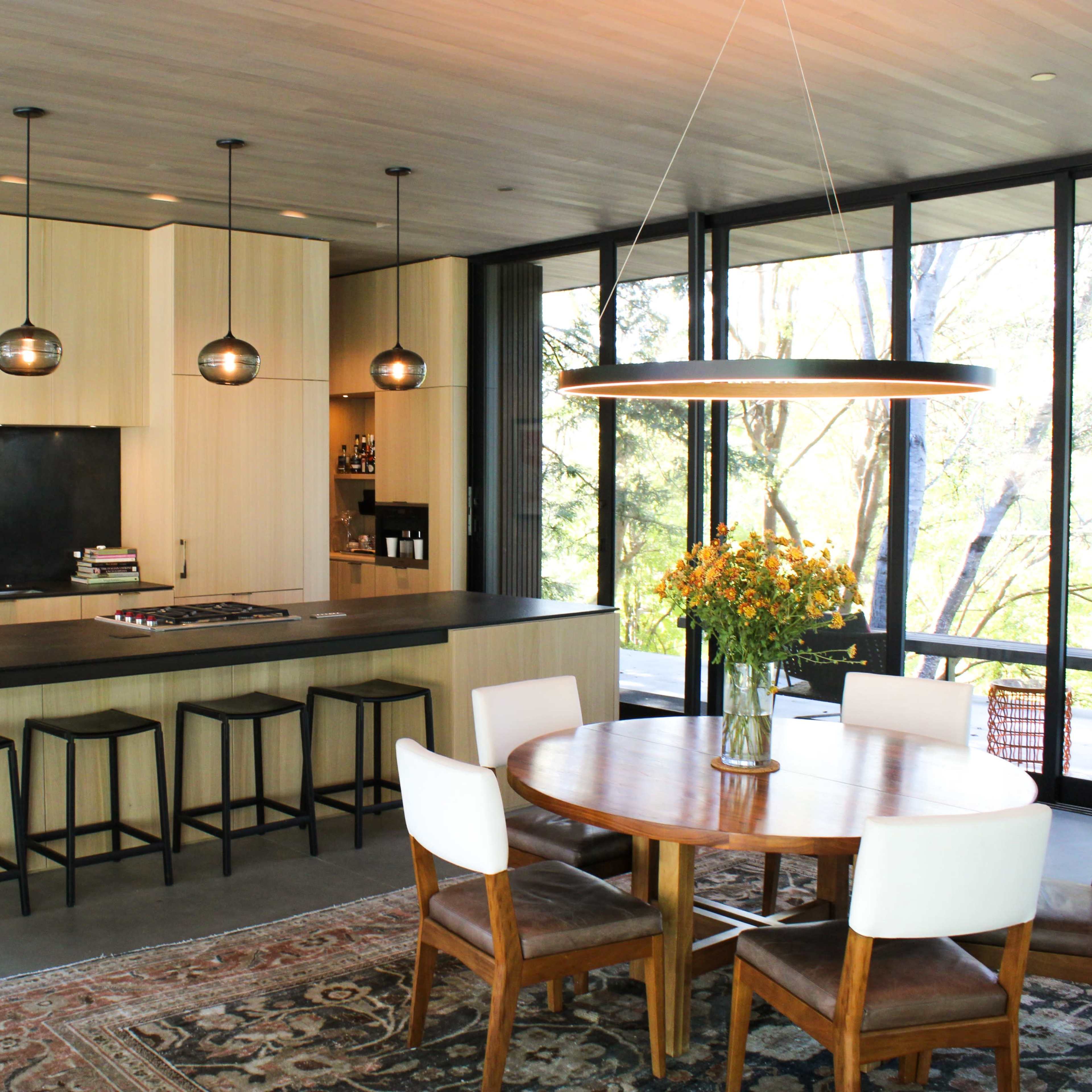 A modern kitchen features a large island with bar stools, sleek cabinetry, and a dining area with a round table and flowers, all illuminated by pendant lights.