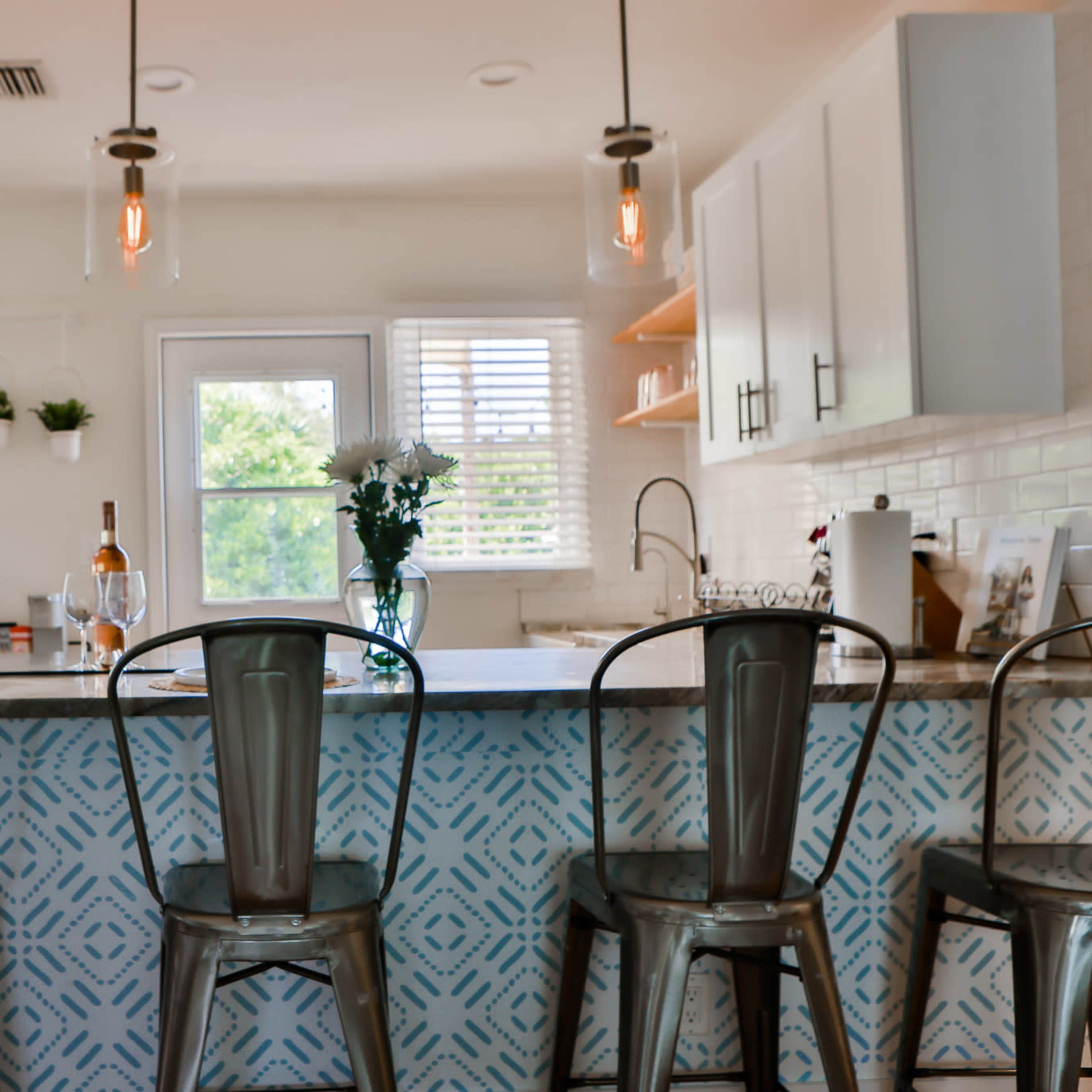 A modern kitchen features a bar with metal stools, a blue-patterned backsplash, and white cabinetry.