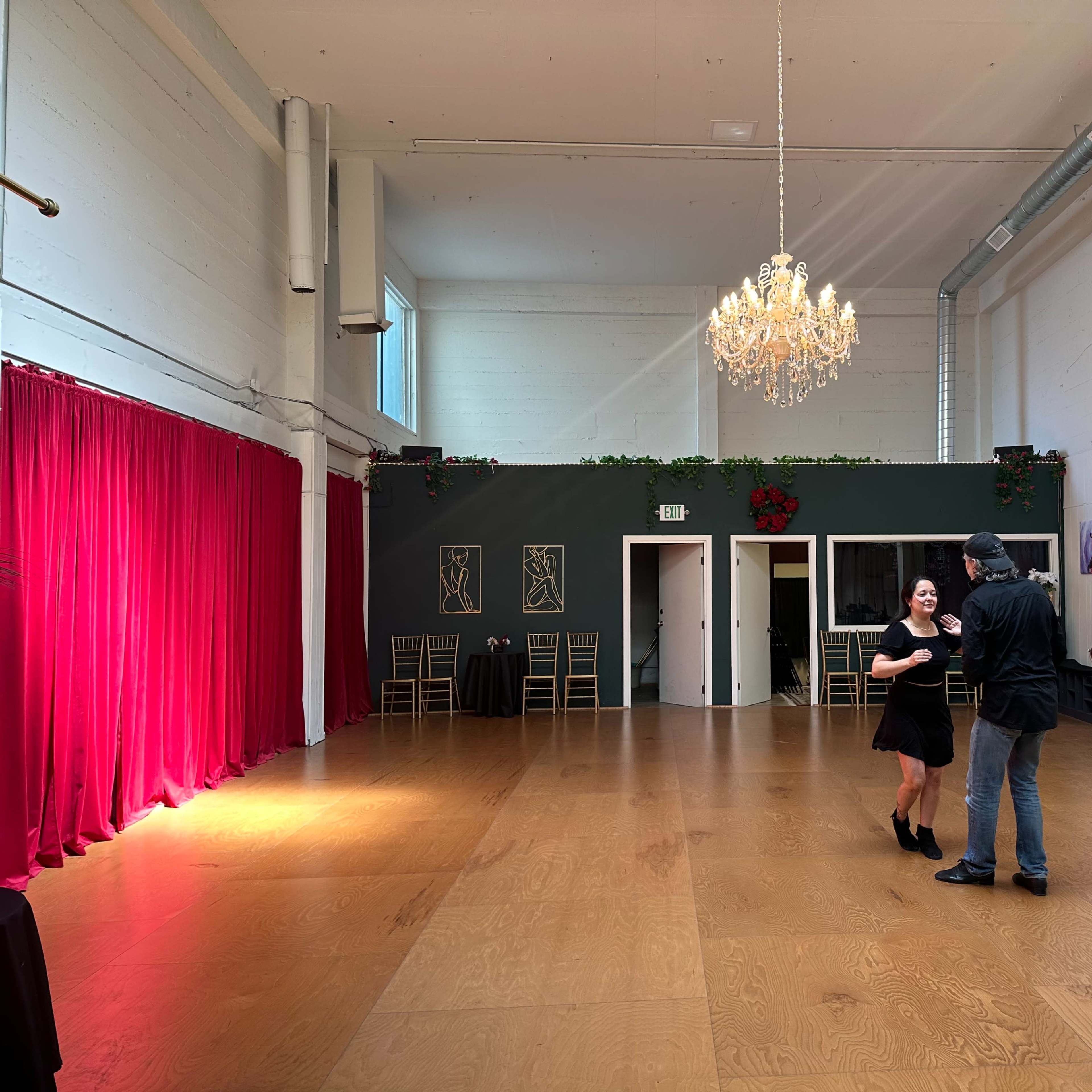 A couple dances in a spacious, well-lit room with a chandelier and red curtains.