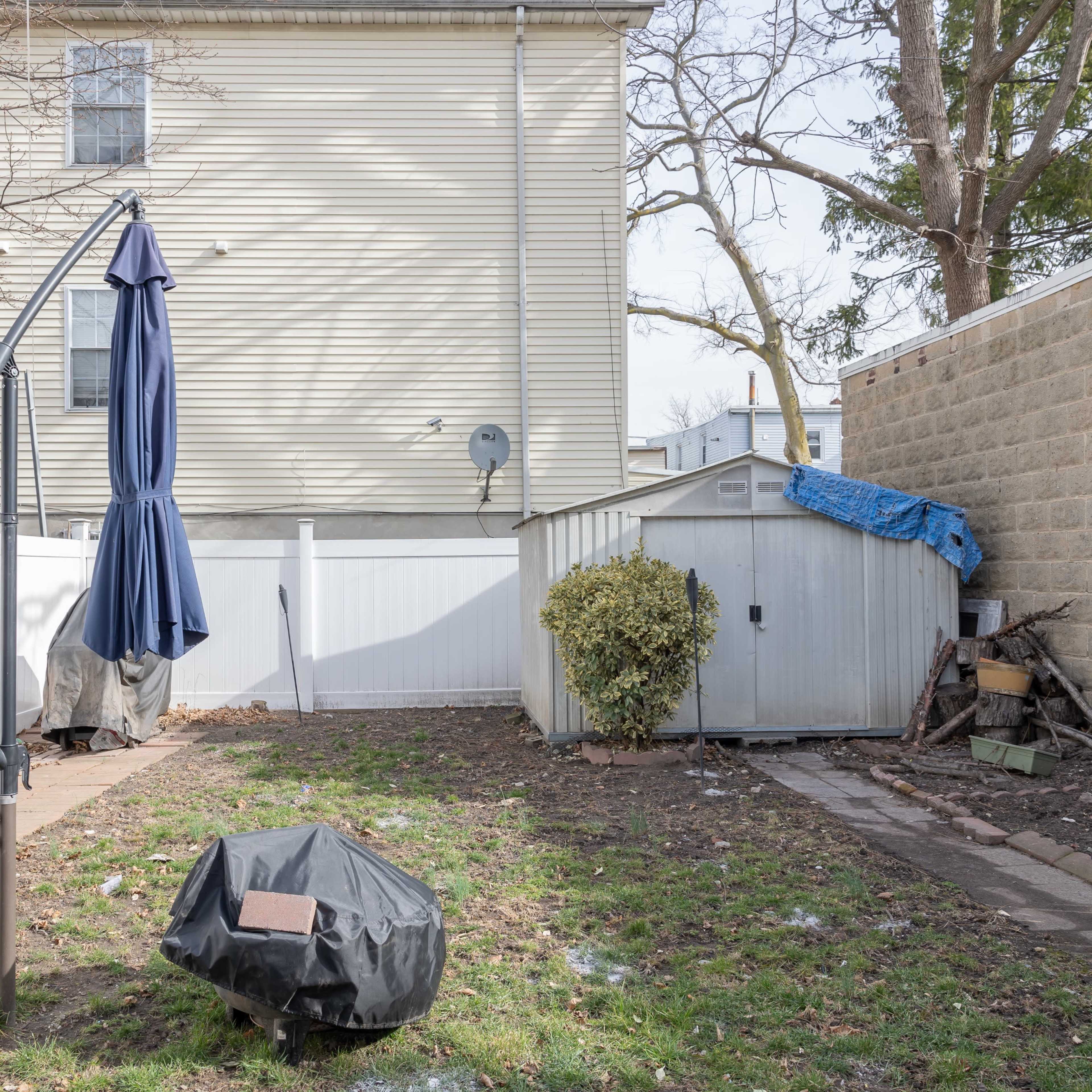 A backyard with a partially collapsed umbrella stand, a shed covered with a blue tarp, and a small patch of grass surrounded by a white fence.