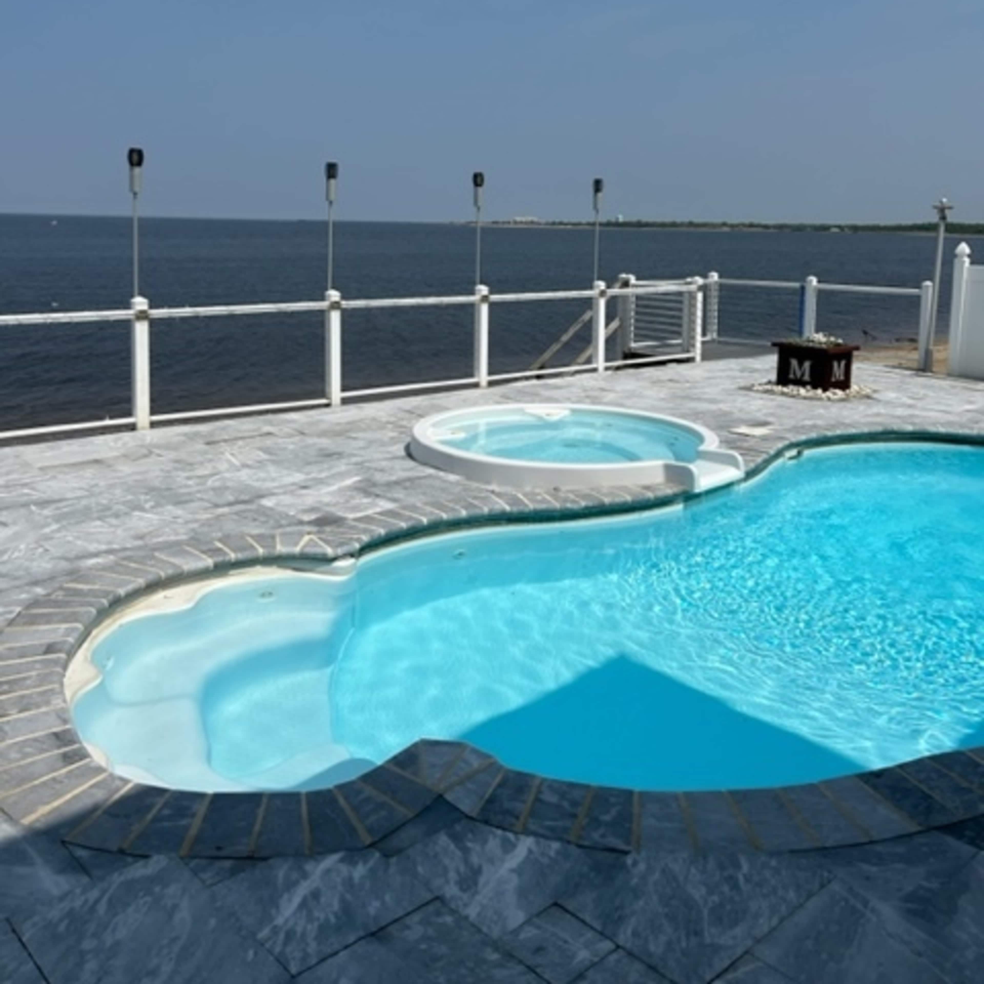 A blue swimming pool with a connected hot tub overlooks a calm body of water under a clear sky.