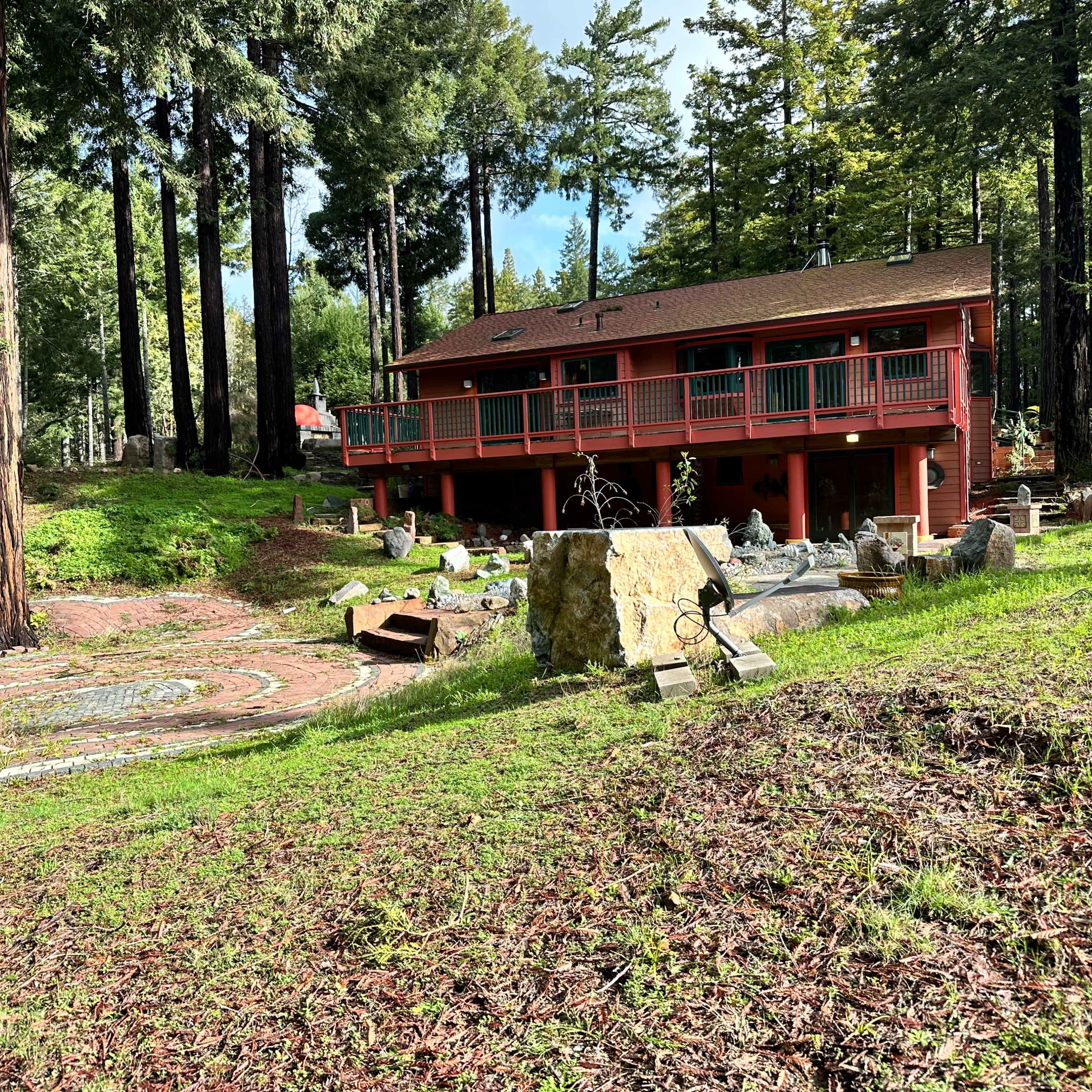 A two-story red house with a balcony is surrounded by tall trees and a landscaped area featuring stones and pathways.