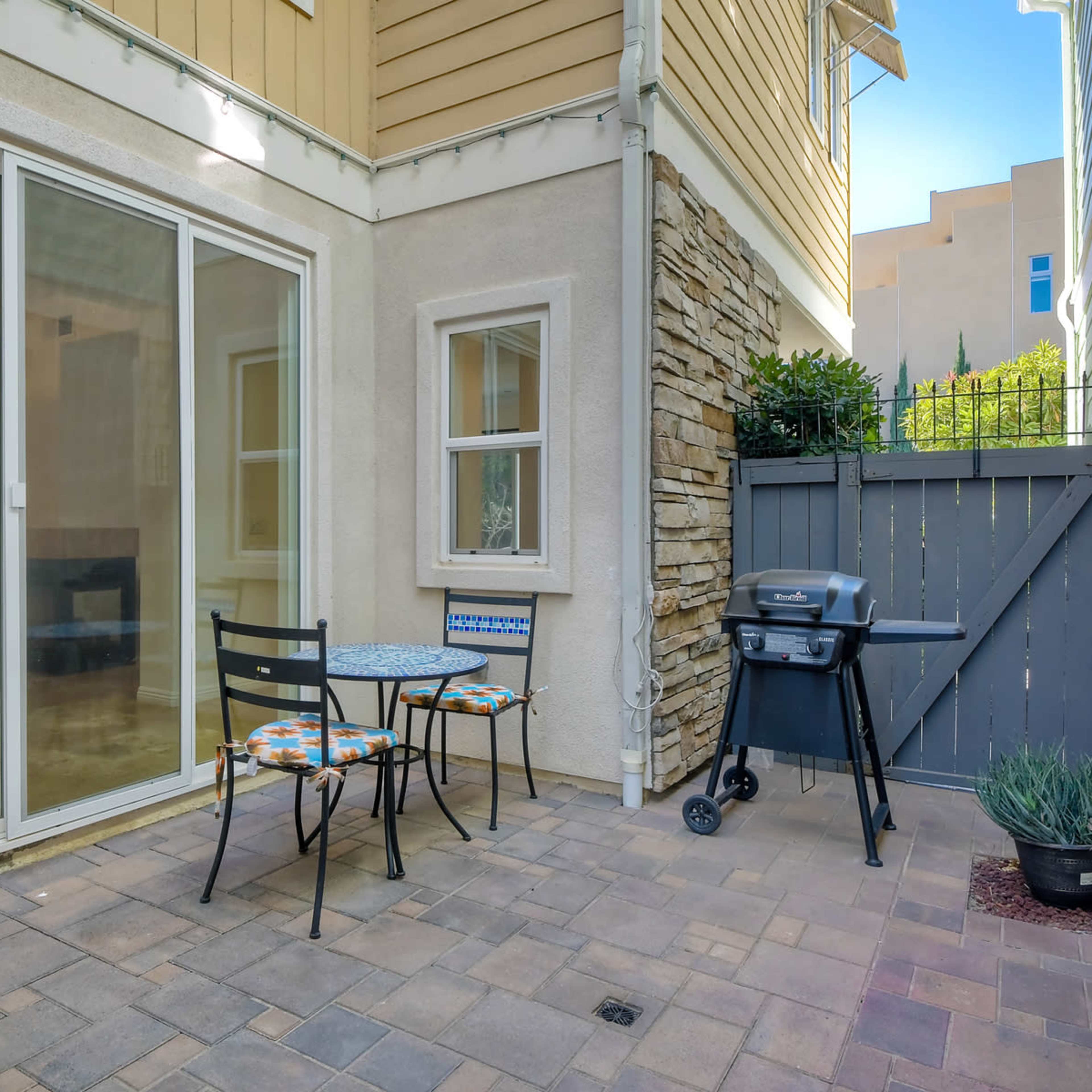 A small outdoor patio features a round table with two chairs and a grill next to a stone wall and sliding glass doors leading into a house.