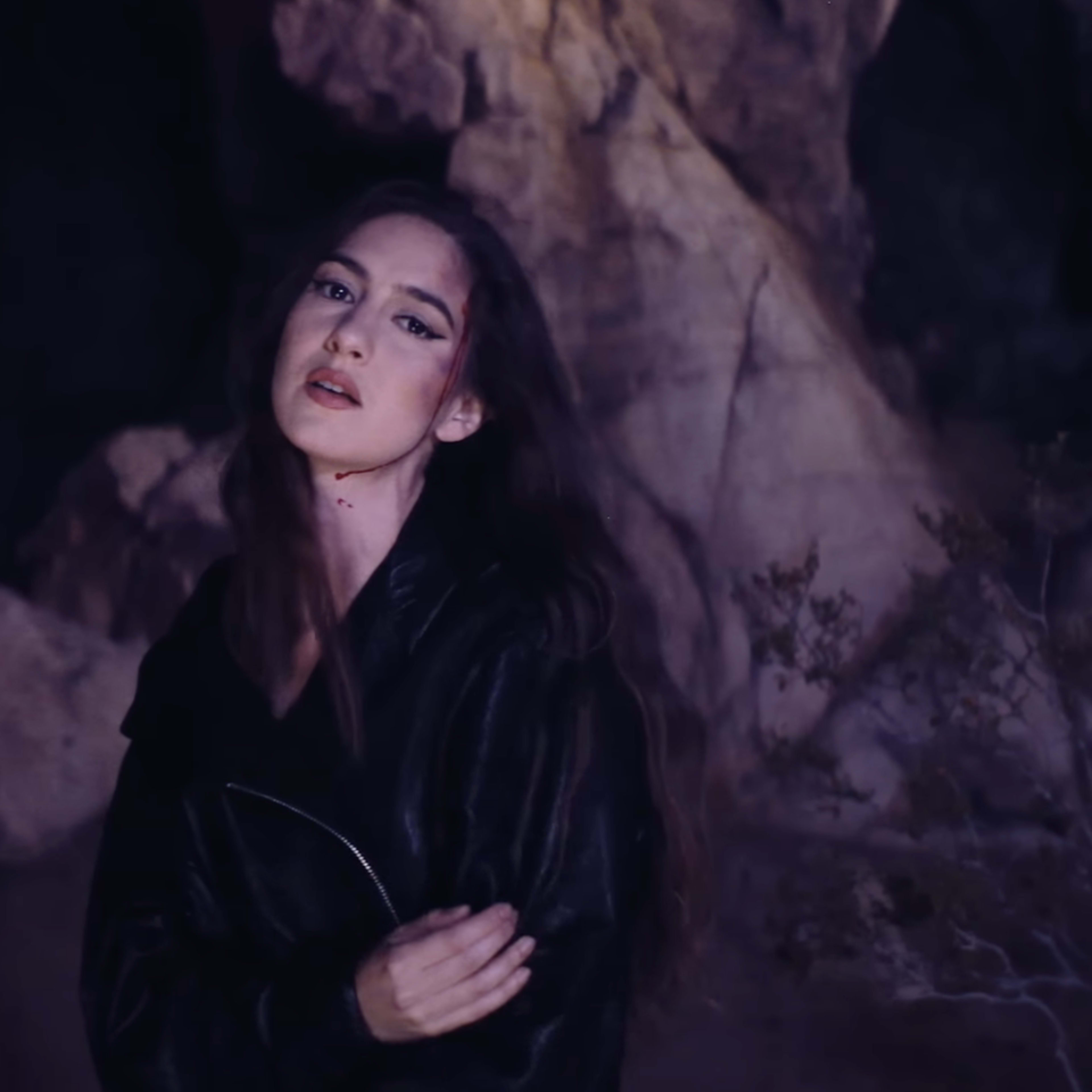 A woman stands in a rocky cave while a shadowy figure with glowing eyes lurks in the background.
