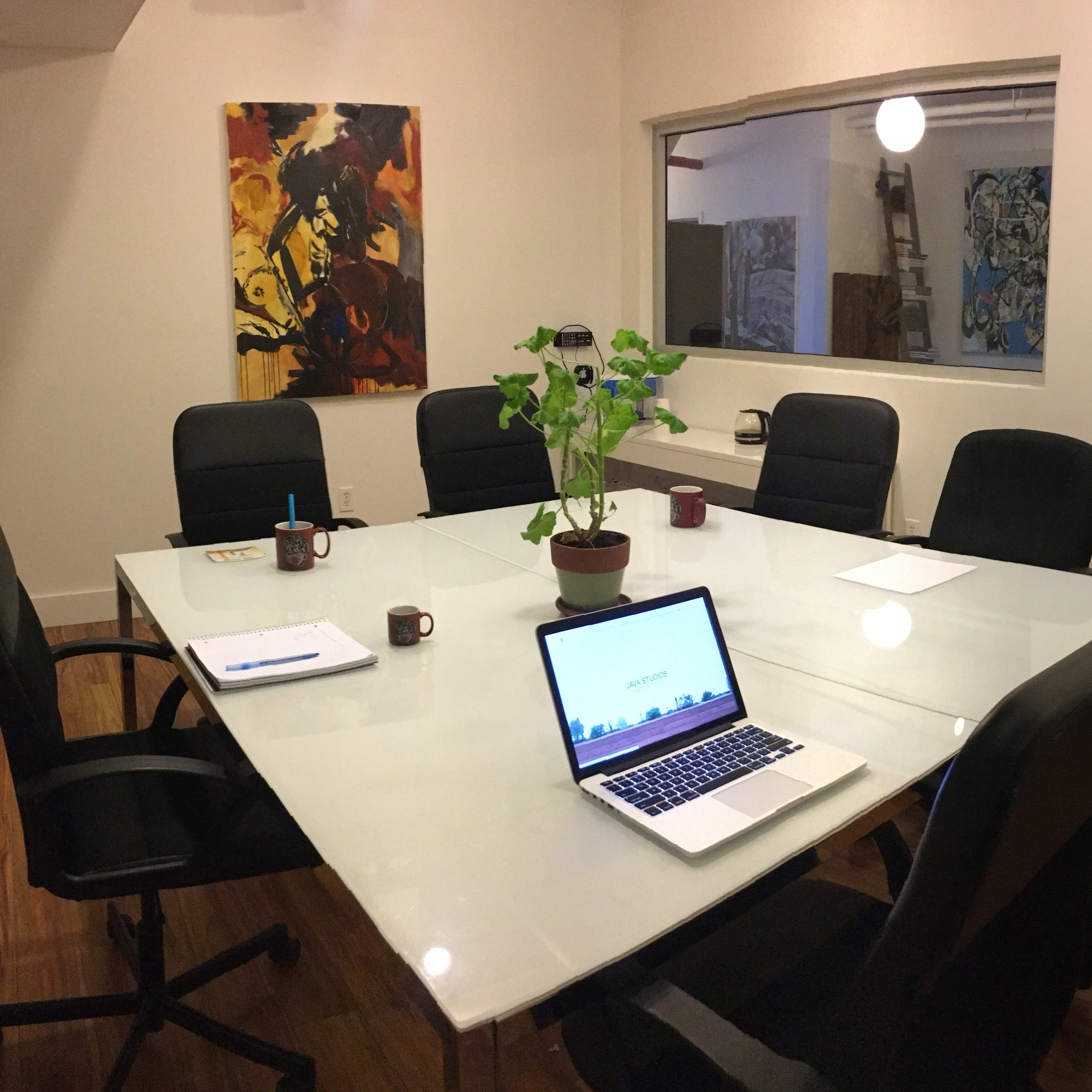 A modern conference room features a glass table surrounded by black chairs, with a laptop open and a potted plant in the center.