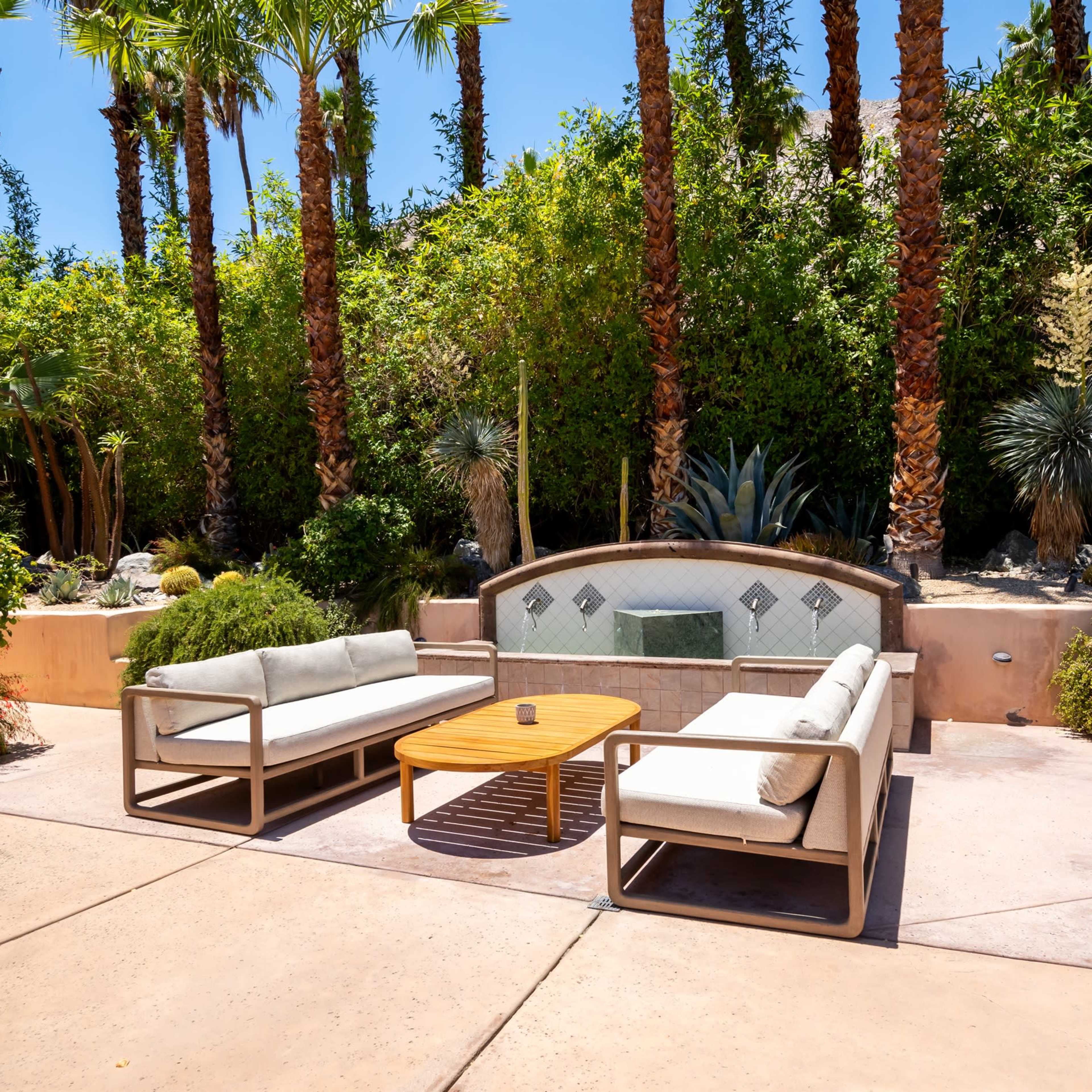 A landscaped outdoor seating area with two couches and a wooden coffee table surrounded by tall palm trees and other greenery.