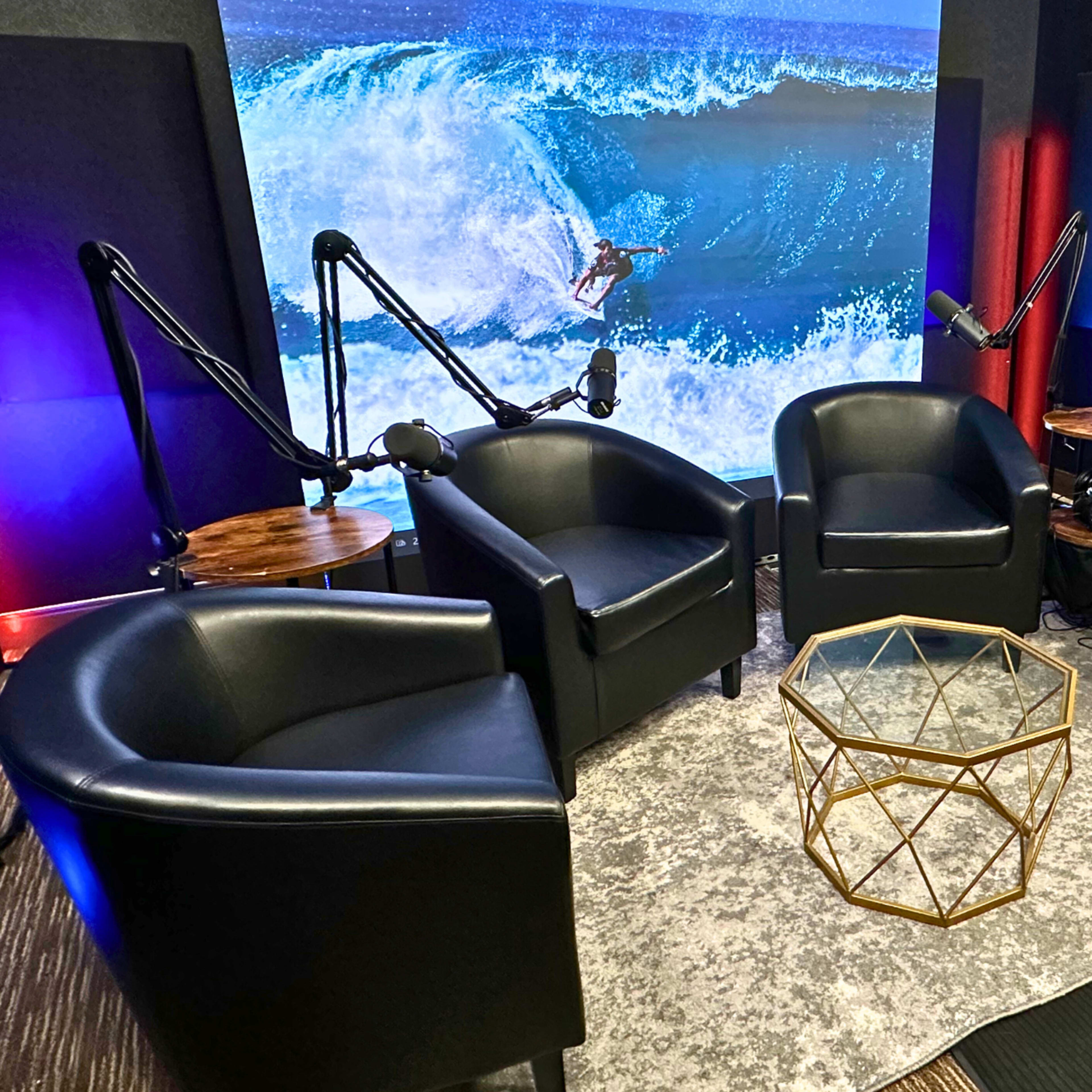 A modern studio setup with four black chairs arranged around a glass coffee table, in front of a large backdrop featuring a surfer riding a wave.