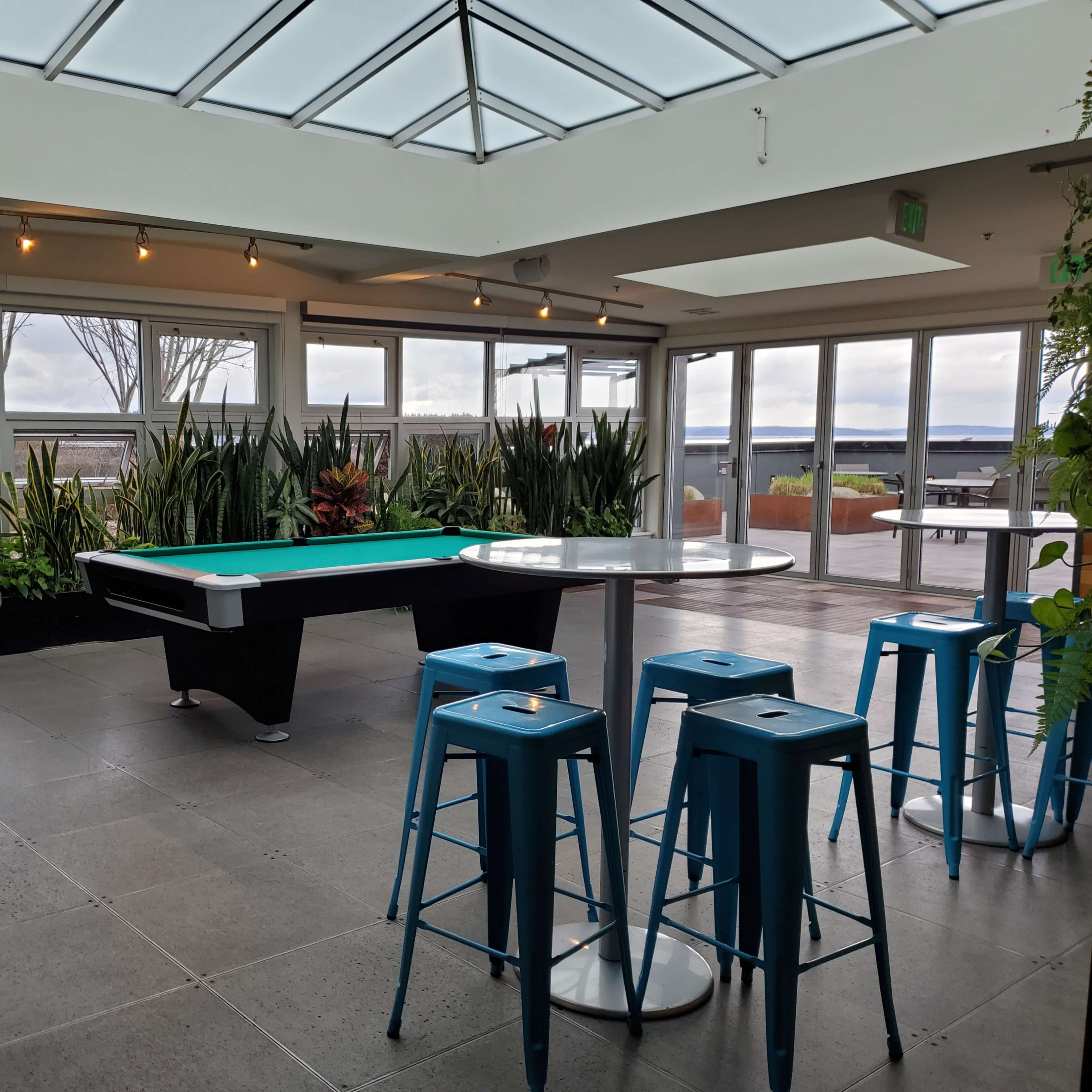 A spacious indoor area featuring a pool table, tall blue stools around a table, and large windows offering a view of plants and an outdoor area.