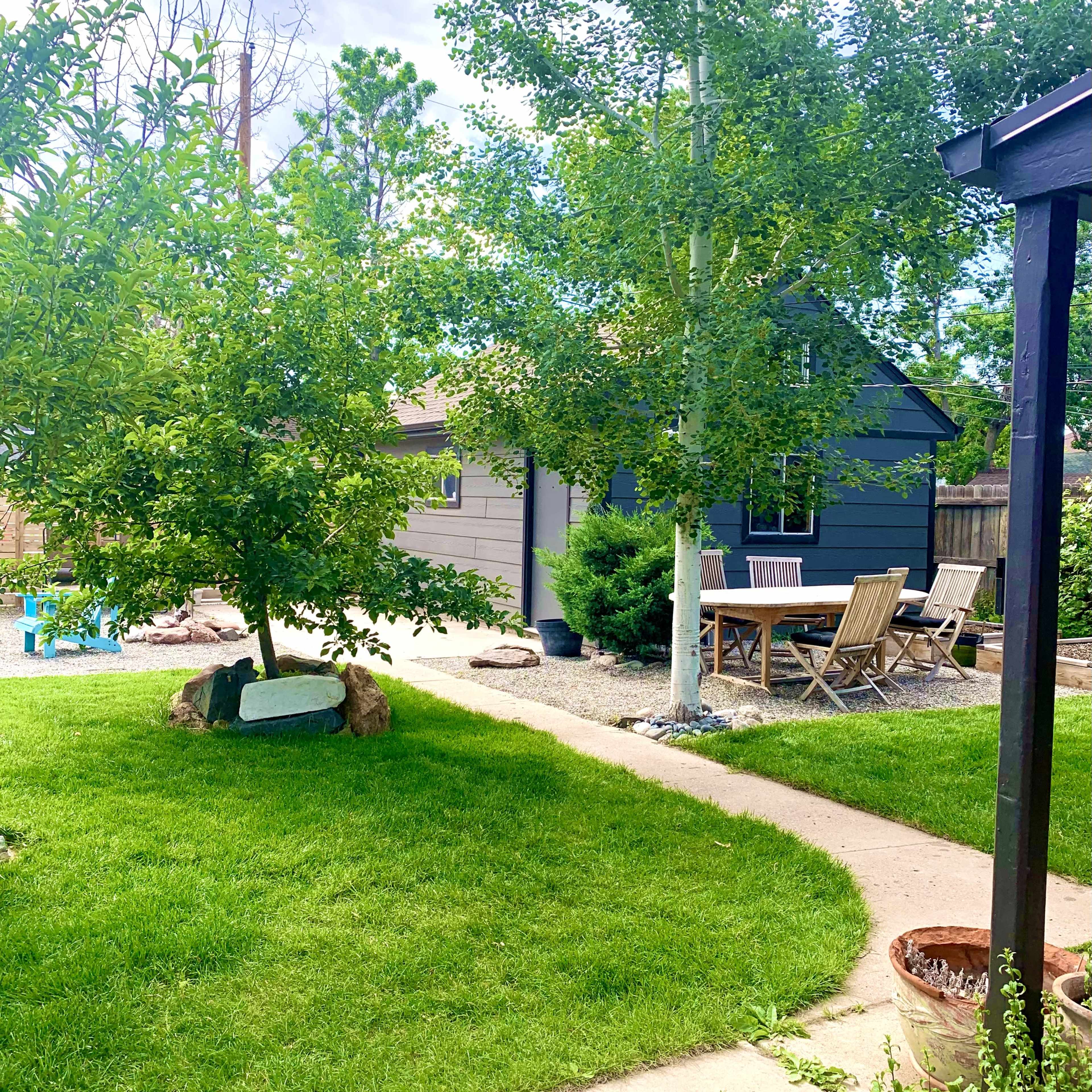 A small, dark blue cottage is situated in a green yard, with a patio set and trees surrounding the area.