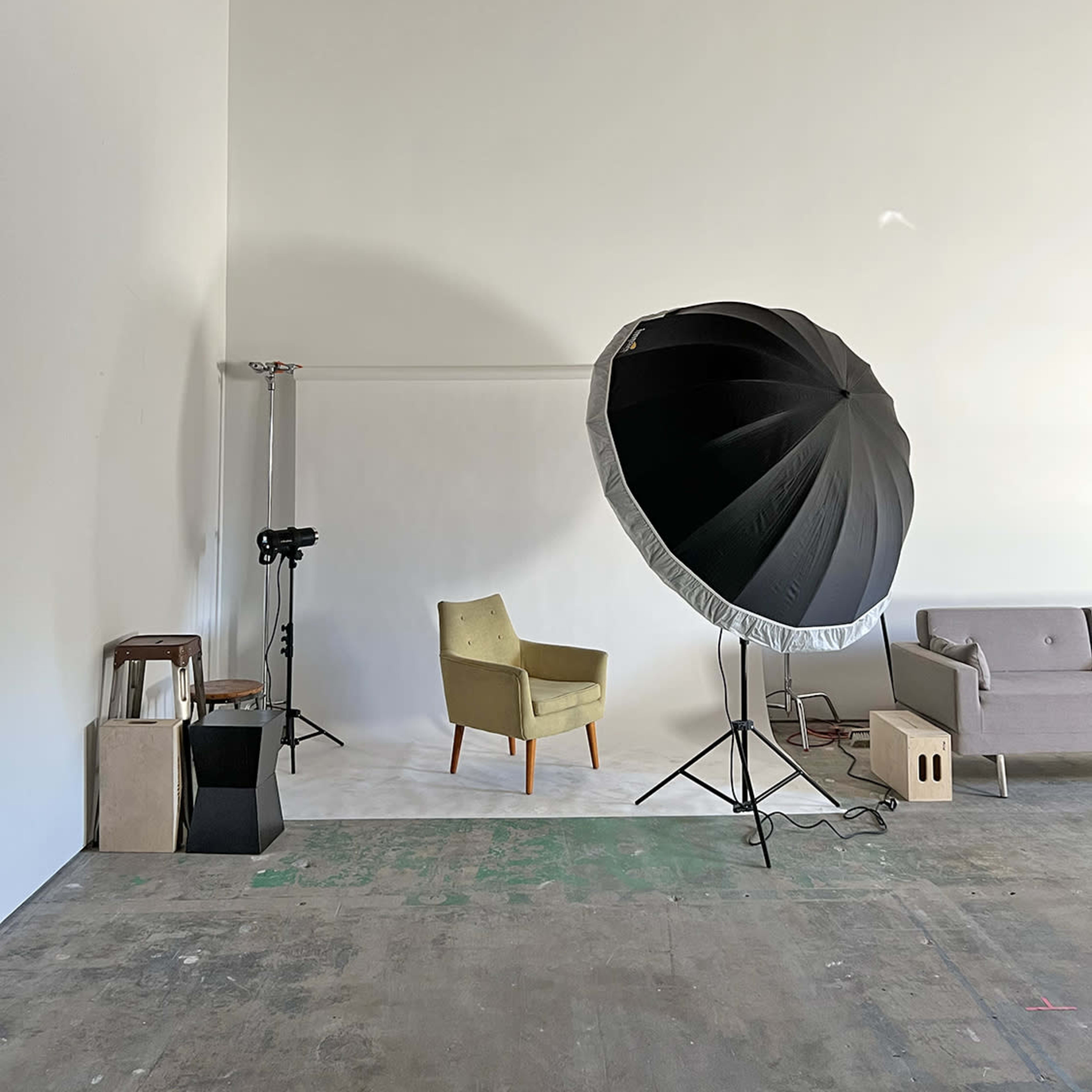 A minimalist studio space featuring a green upholstered chair, a gray couch, a large softbox light, and various props arranged against a white wall.