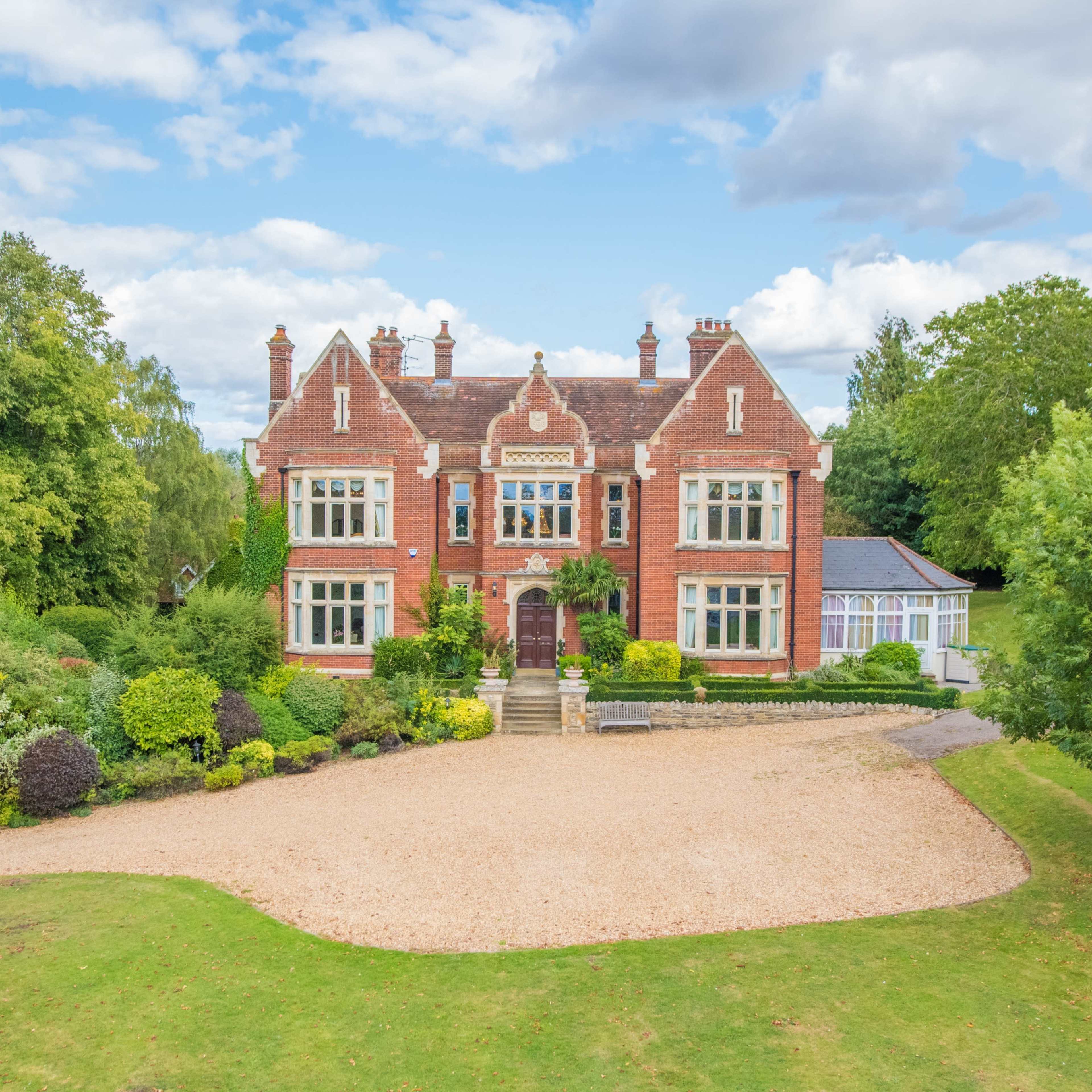 A large red brick mansion is surrounded by well-maintained gardens and trees, set against a partly cloudy sky.