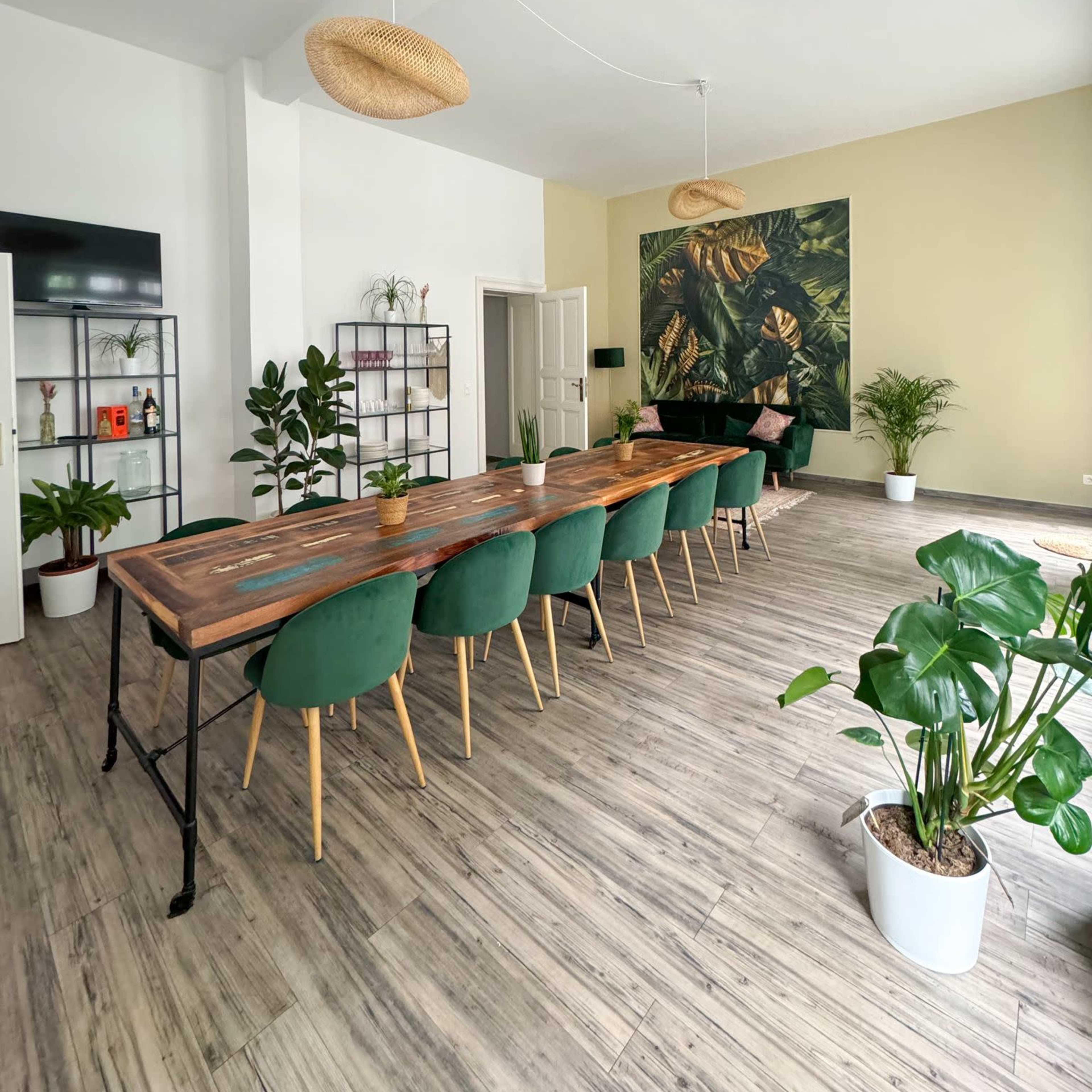 A spacious dining area features a long wooden table surrounded by green chairs, potted plants, and decorative wall art with tropical leaves.
