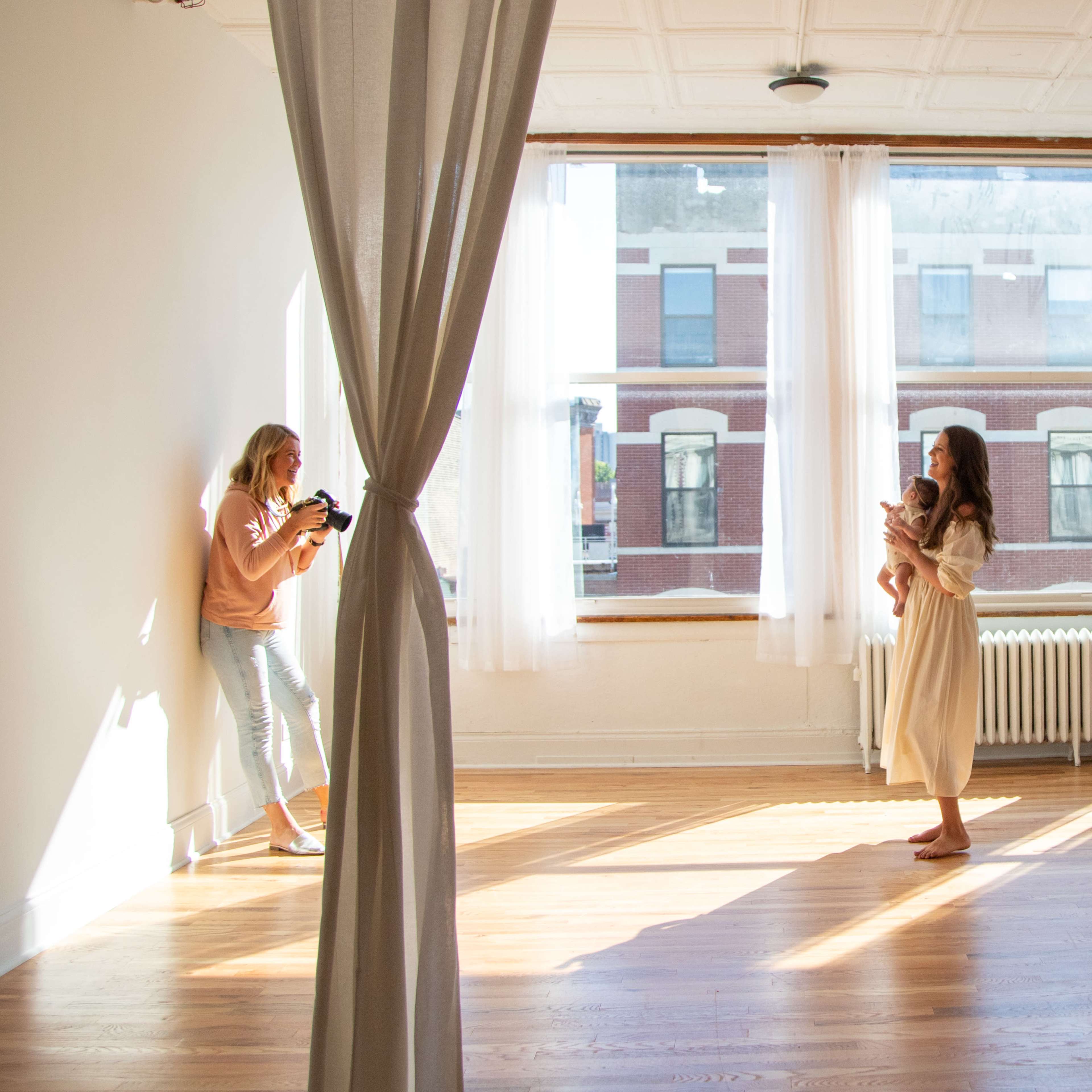 A woman photographs another woman holding a baby in a sunlit, spacious room with large windows.