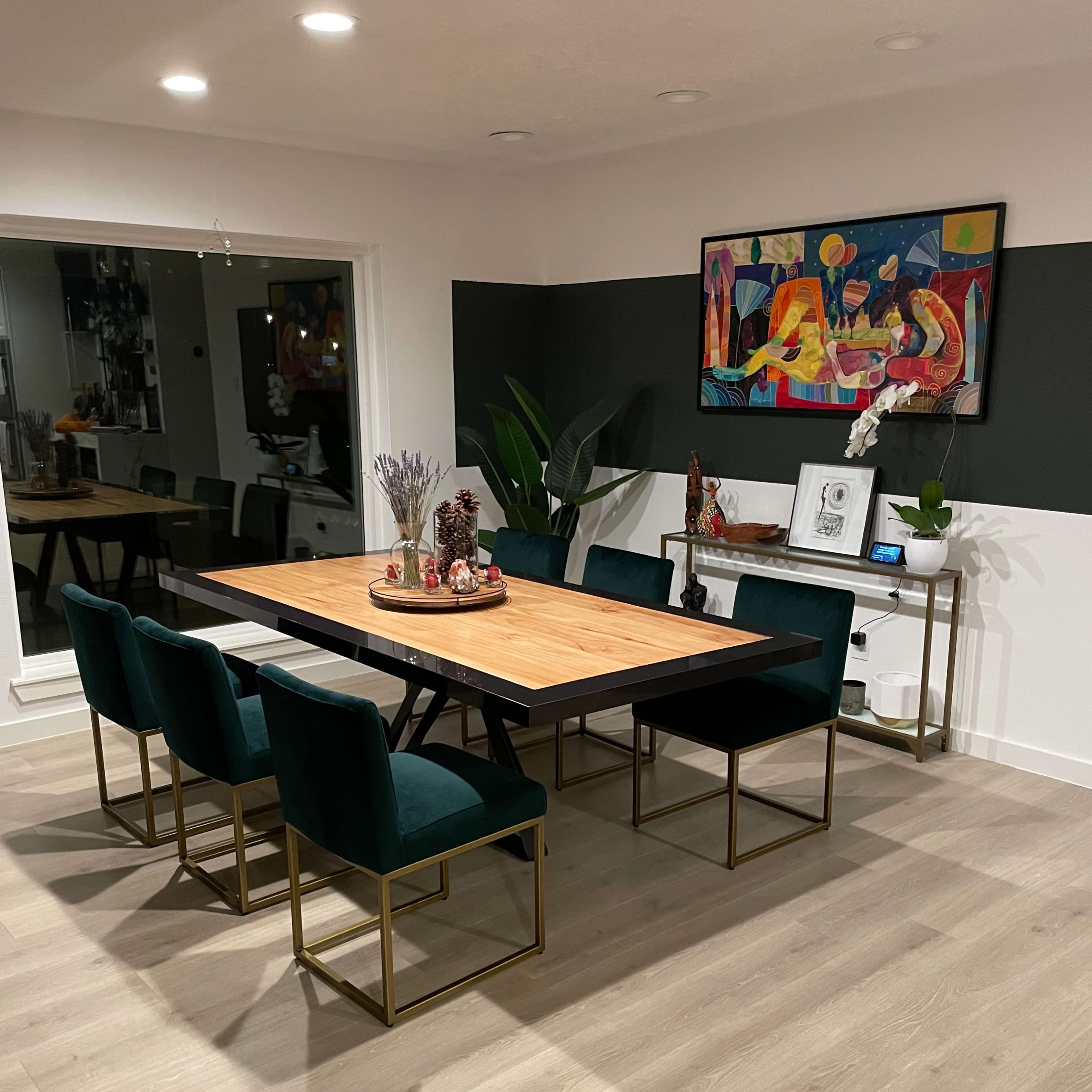 A modern dining room features a wooden table surrounded by six green chairs, with a large artwork hanging on the wall and a console table displayed against a contrasting black backdrop.