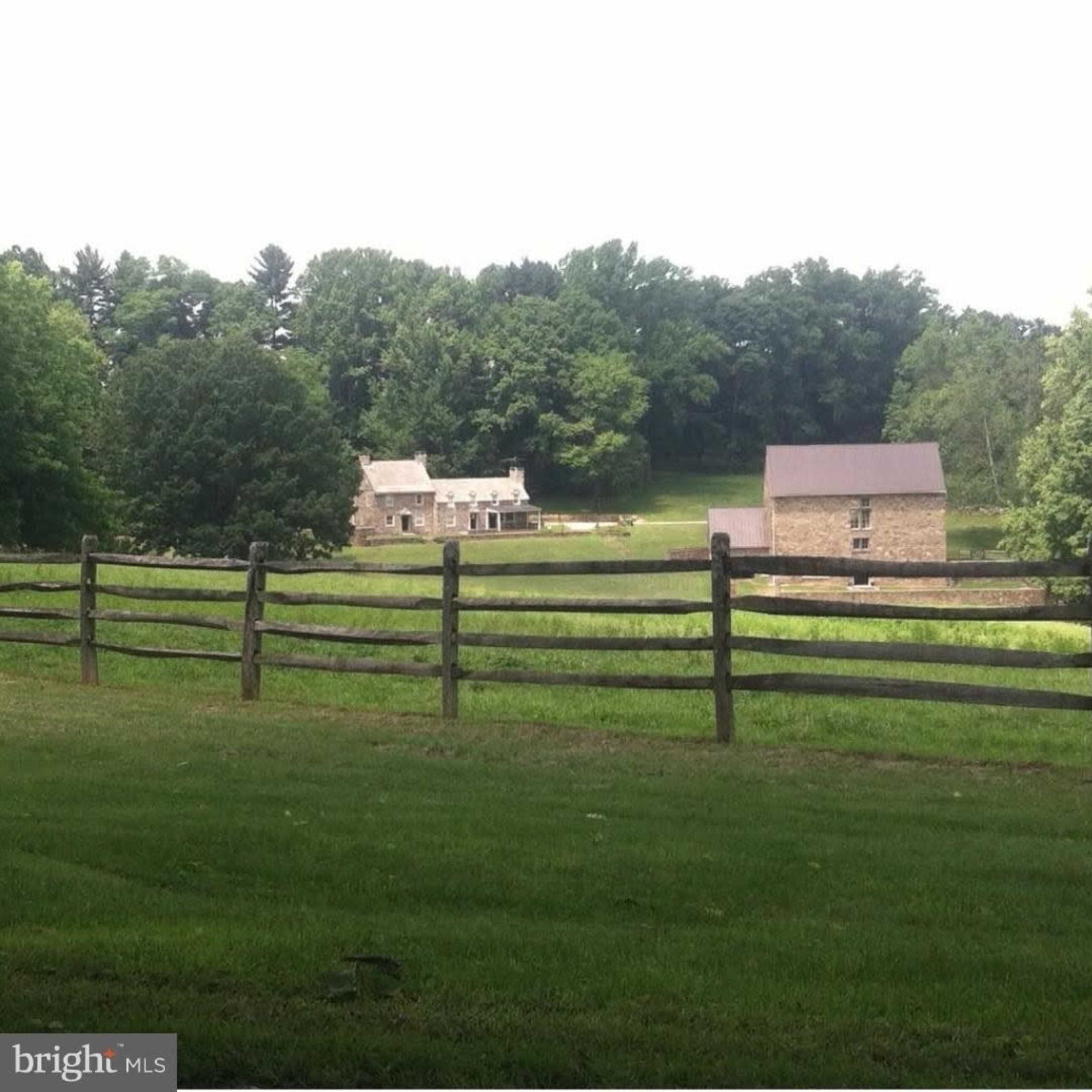 A rural landscape with a wooden fence in the foreground and two stone buildings set against a backdrop of trees and open fields.